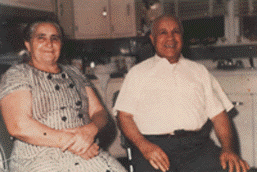 Two people sitting in a kitchen, smiling, with cabinets and appliances in the background.