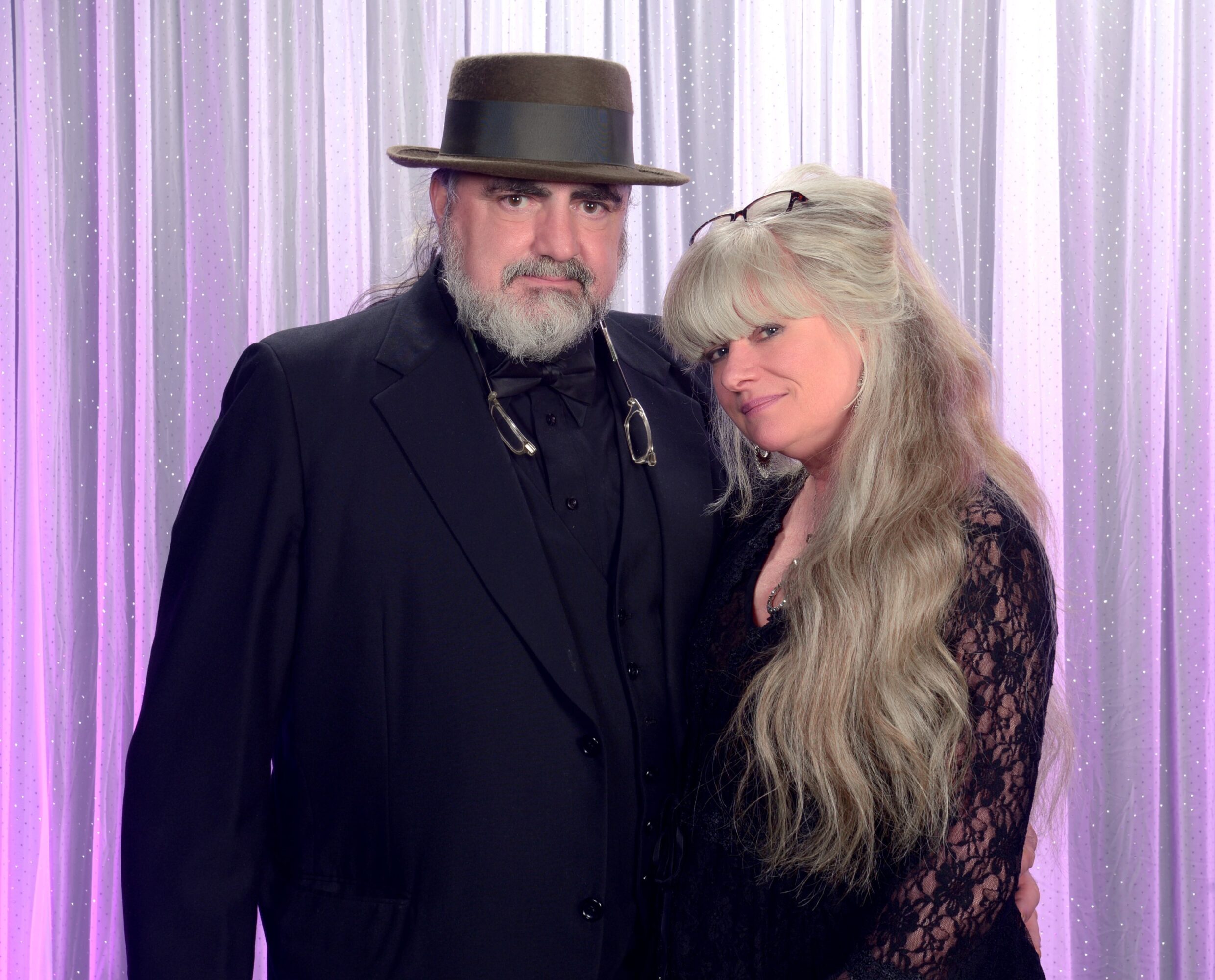 A man and woman pose together in front of a purple curtain backdrop, both dressed in dark clothing, with the woman having long, wavy hair.