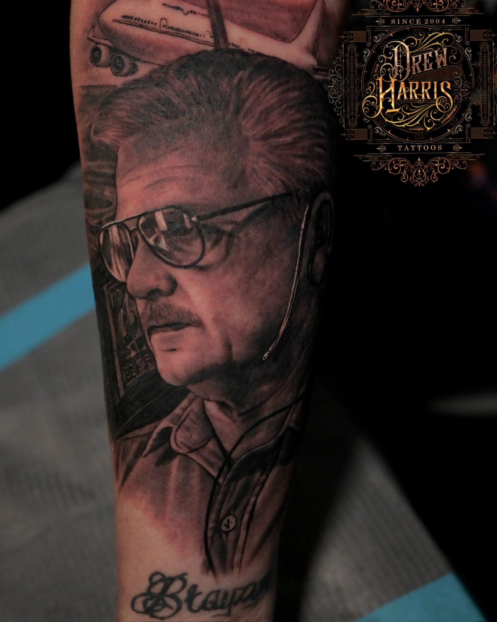 Tattoo of an older man with glasses and a mustache, wearing a collared shirt, on a person's arm.