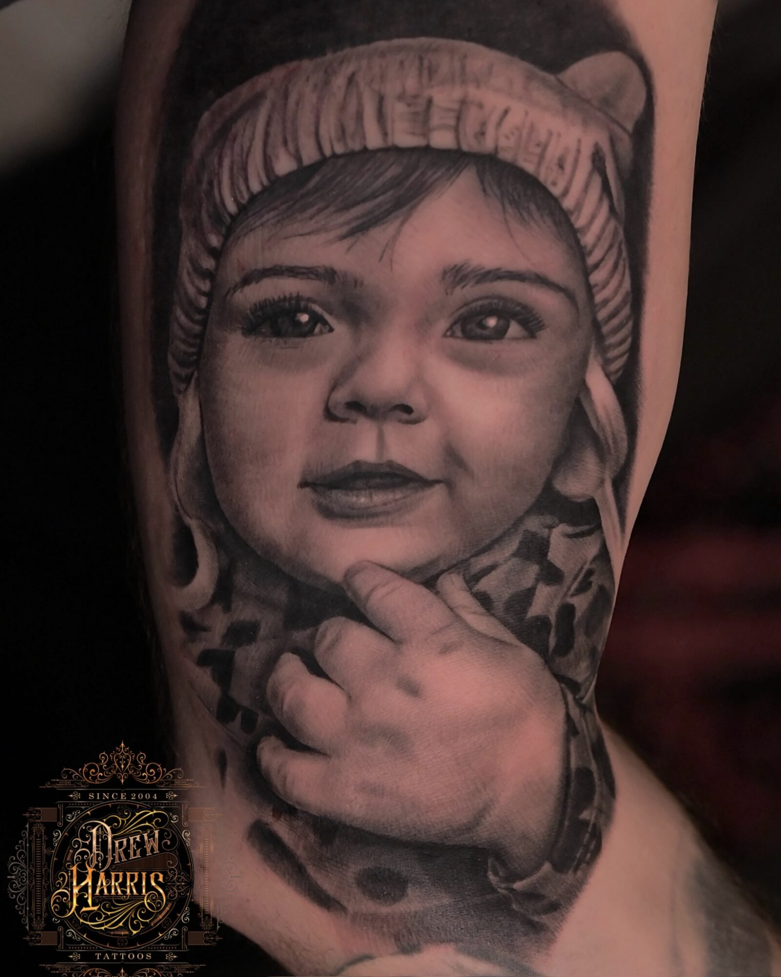 Realistic black and gray tattoo of a young child's face wearing a beanie, with hand near chin, detailed shading.