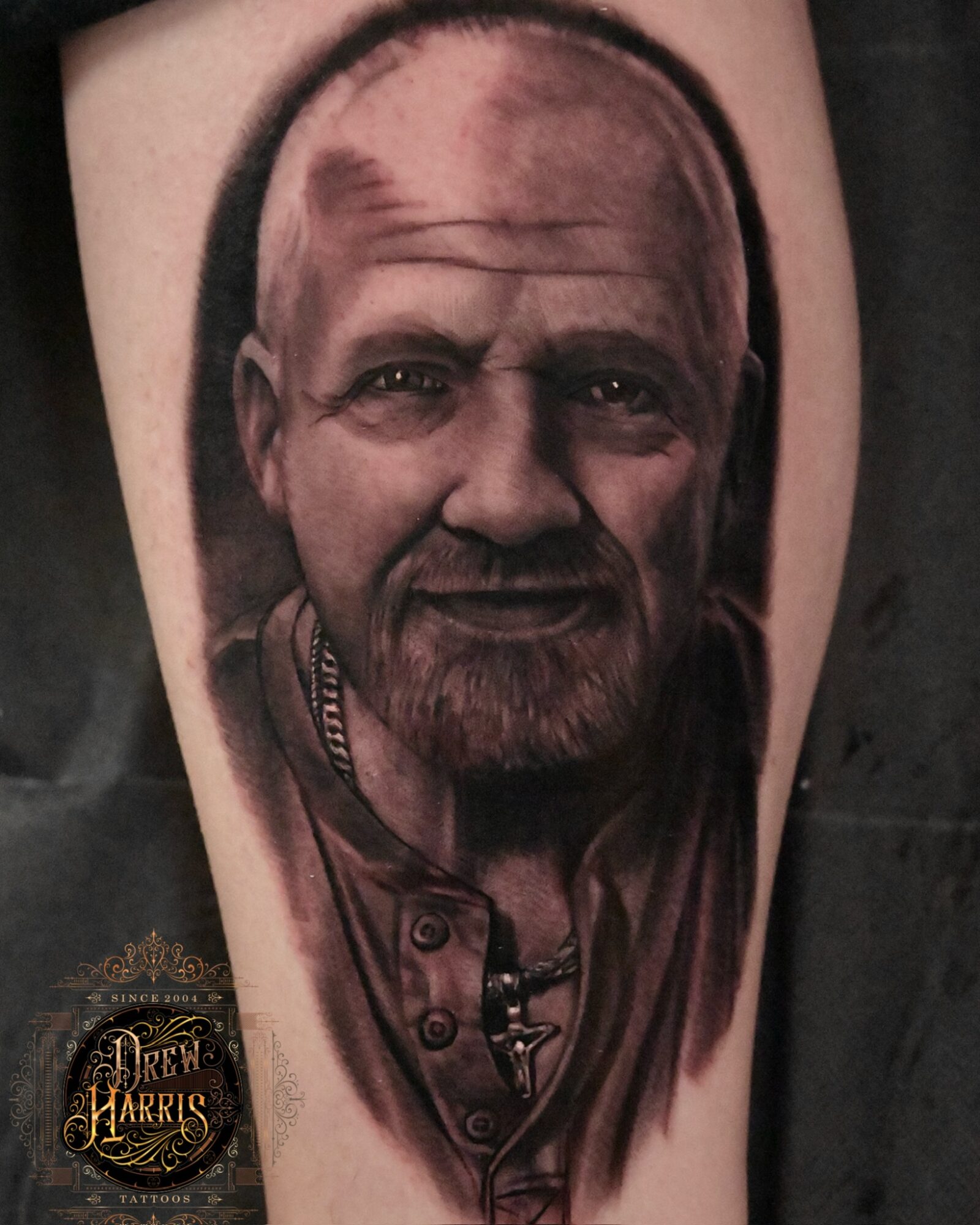 Tattoo of a man's face with a beard and bald head, detailed shading, wearing a jacket and necklace, on skin.