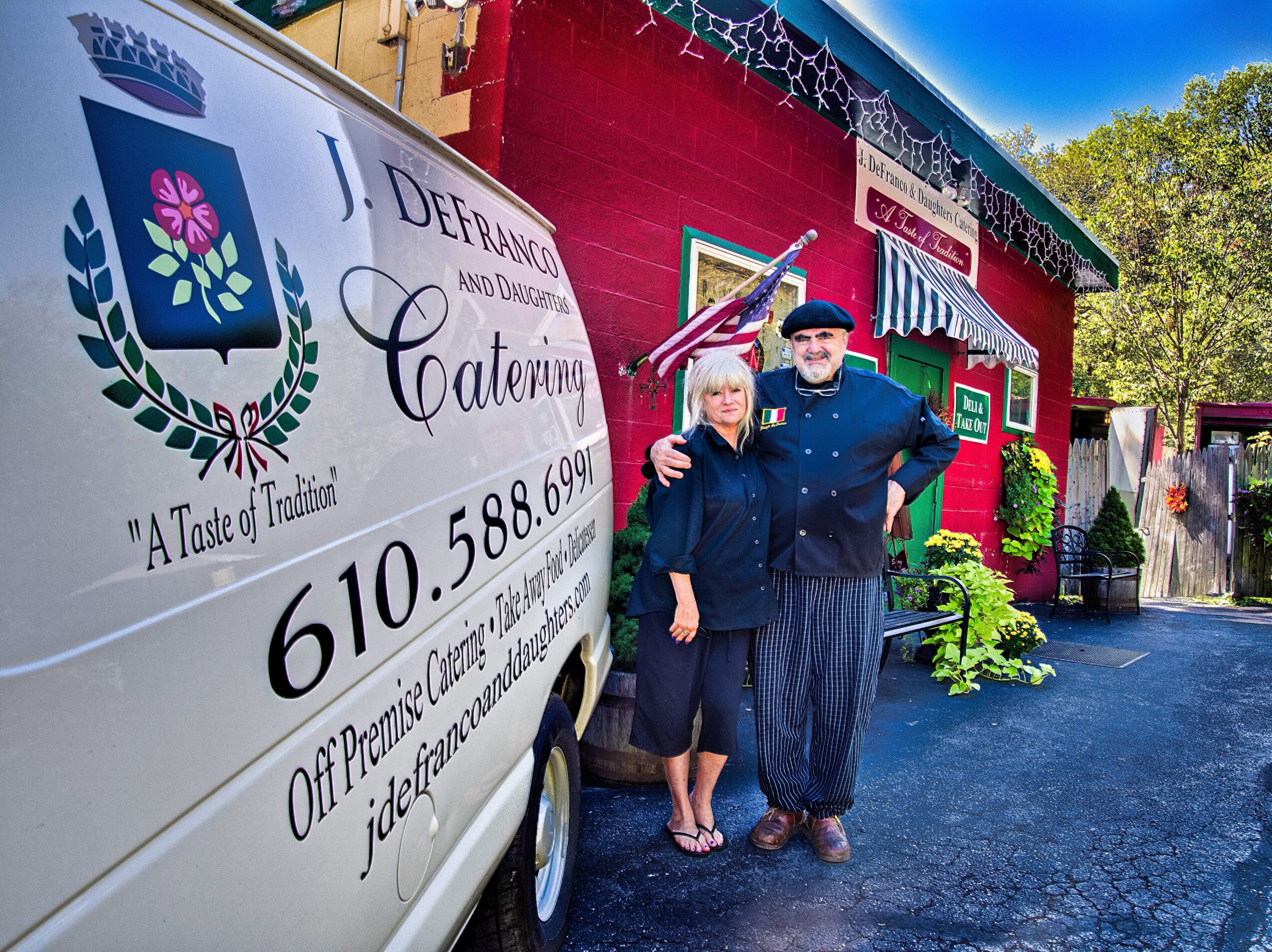 Two people stand outside a red building with greenery, next to a white van with a logo and contact information.