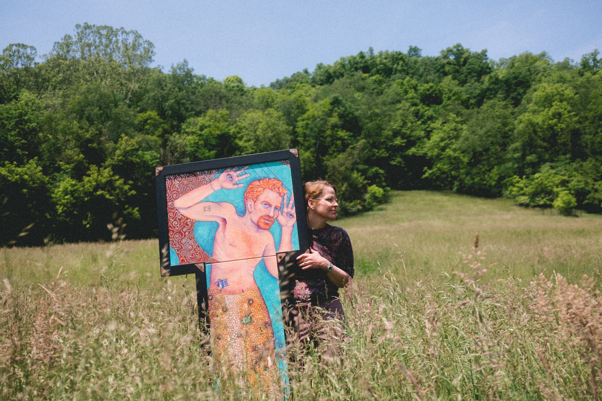 Person standing in a grassy field with a colorful painting on a board, surrounded by trees and blue sky.