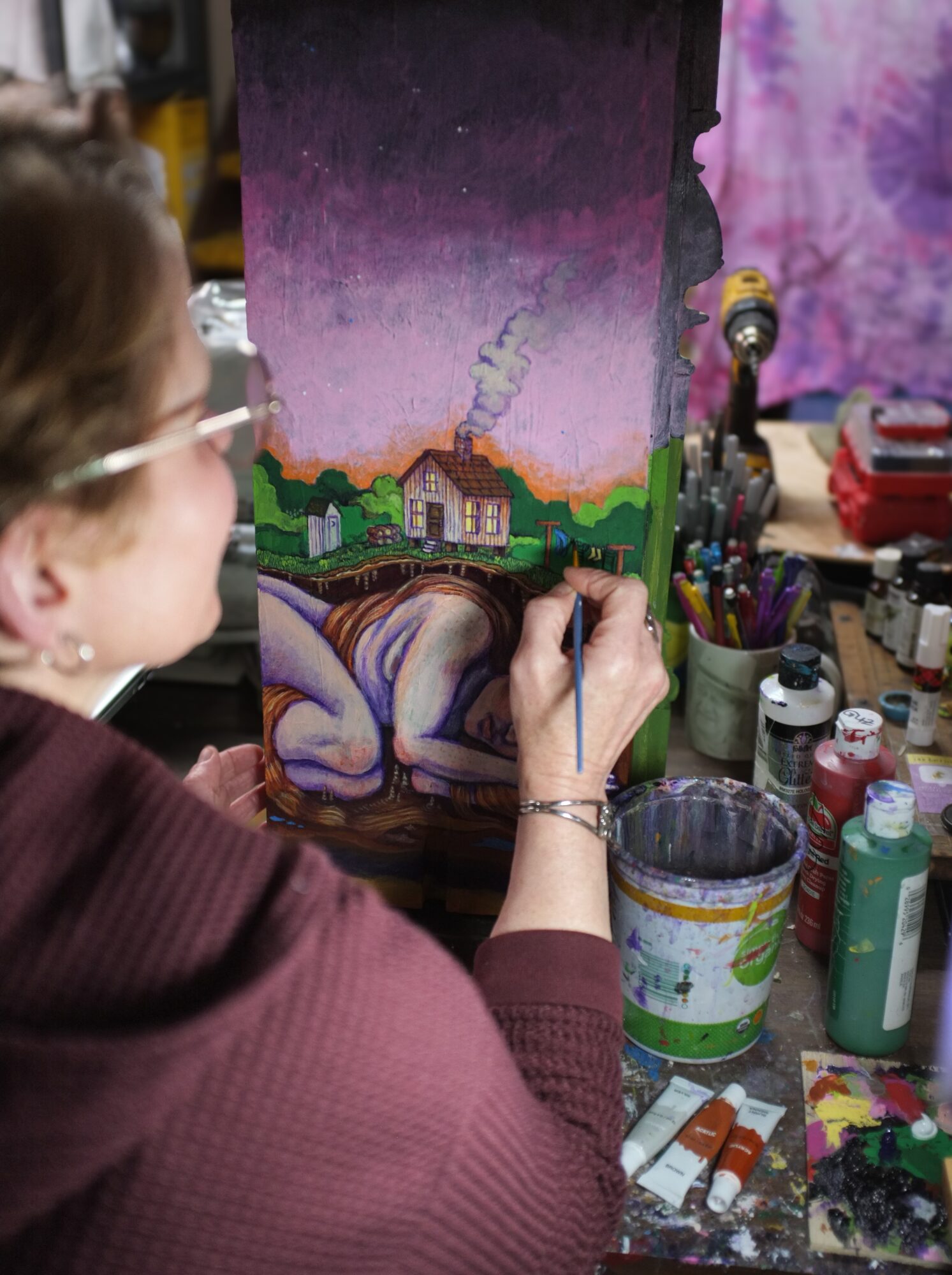 Person painting a colorful landscape with a house, trees, and a sky with stars, surrounded by art supplies.