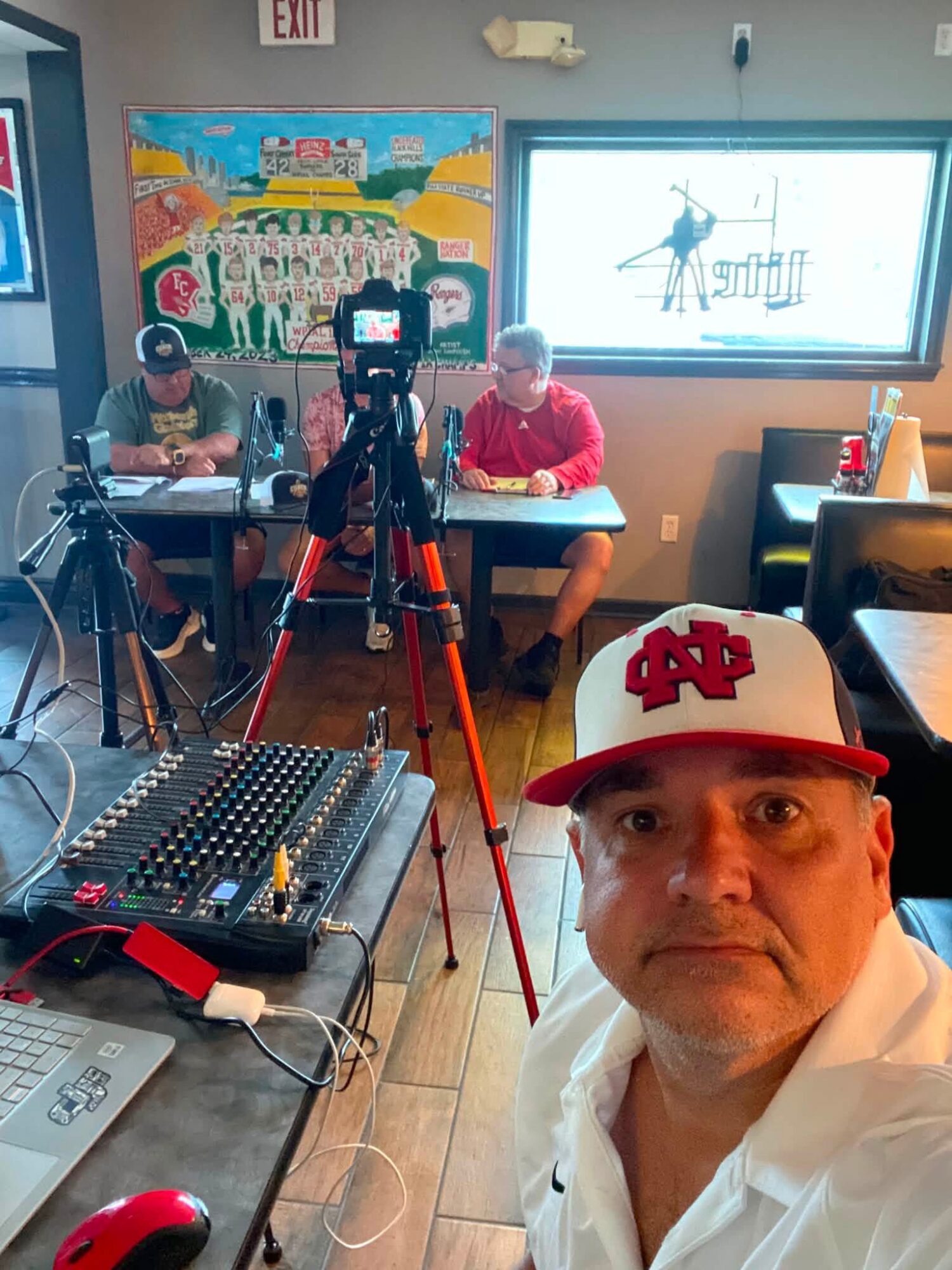 Man wearing a red and white cap takes a selfie in a cafe with two people at a table, camera on tripod, and equipment visible.