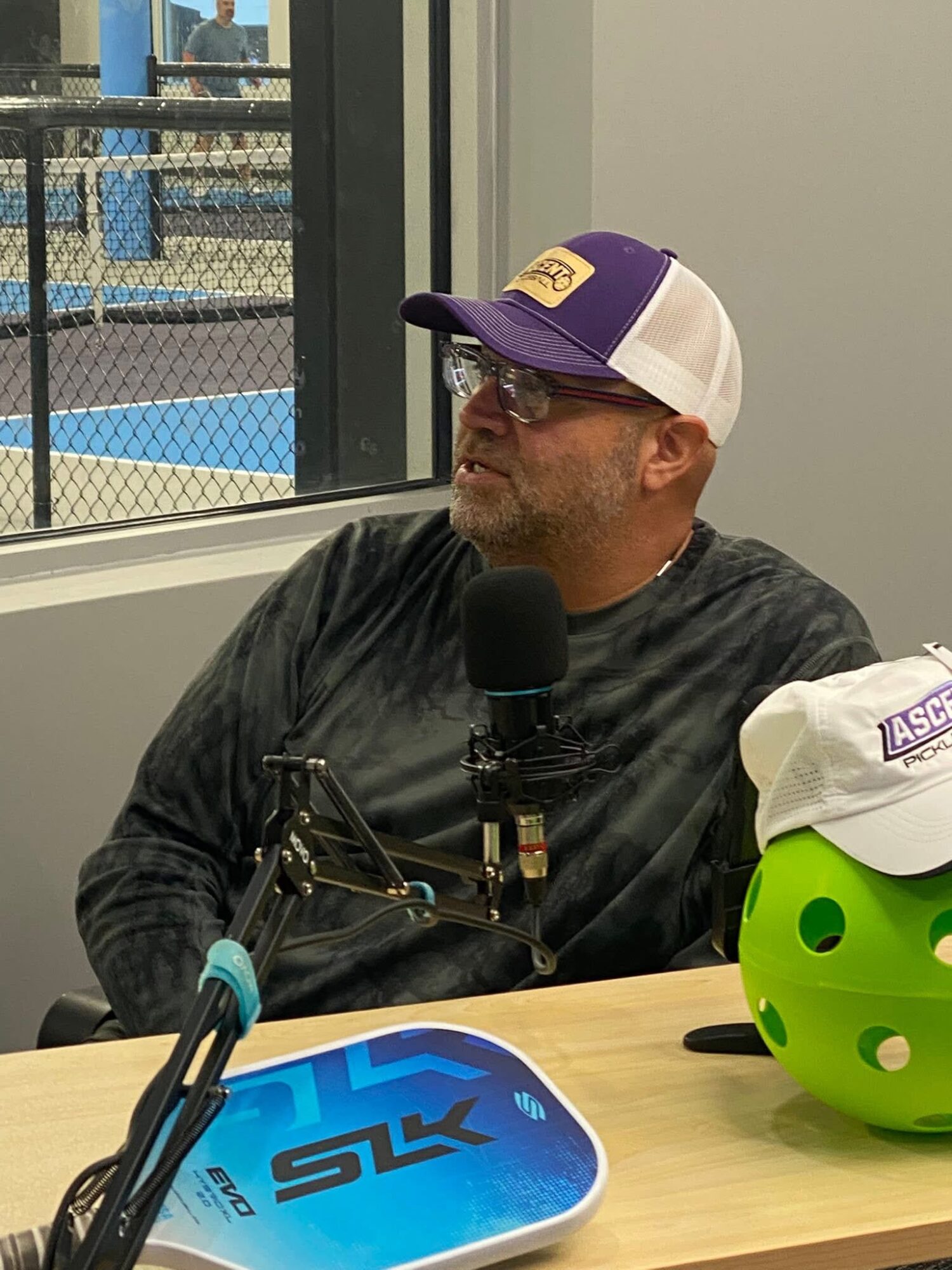Man wearing a purple and white cap, glasses, and a dark shirt, speaking into a microphone, with a green helmet and a blue skateboard on table.