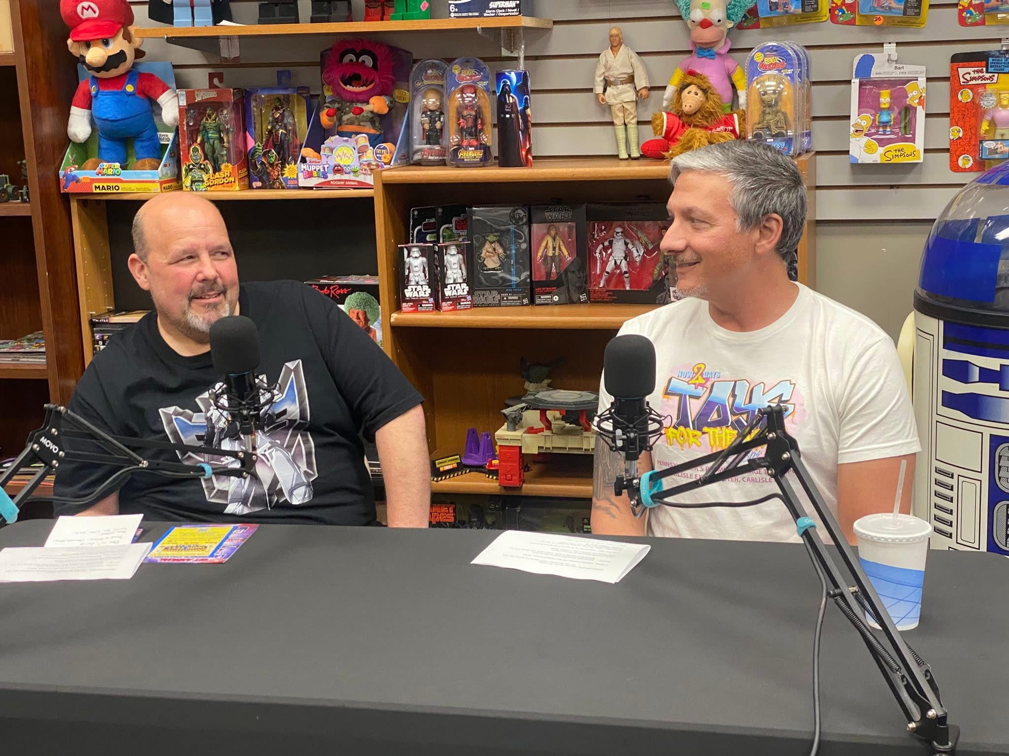 Two men sit at a table with microphones, smiling and talking, surrounded by shelves of toys and collectibles.