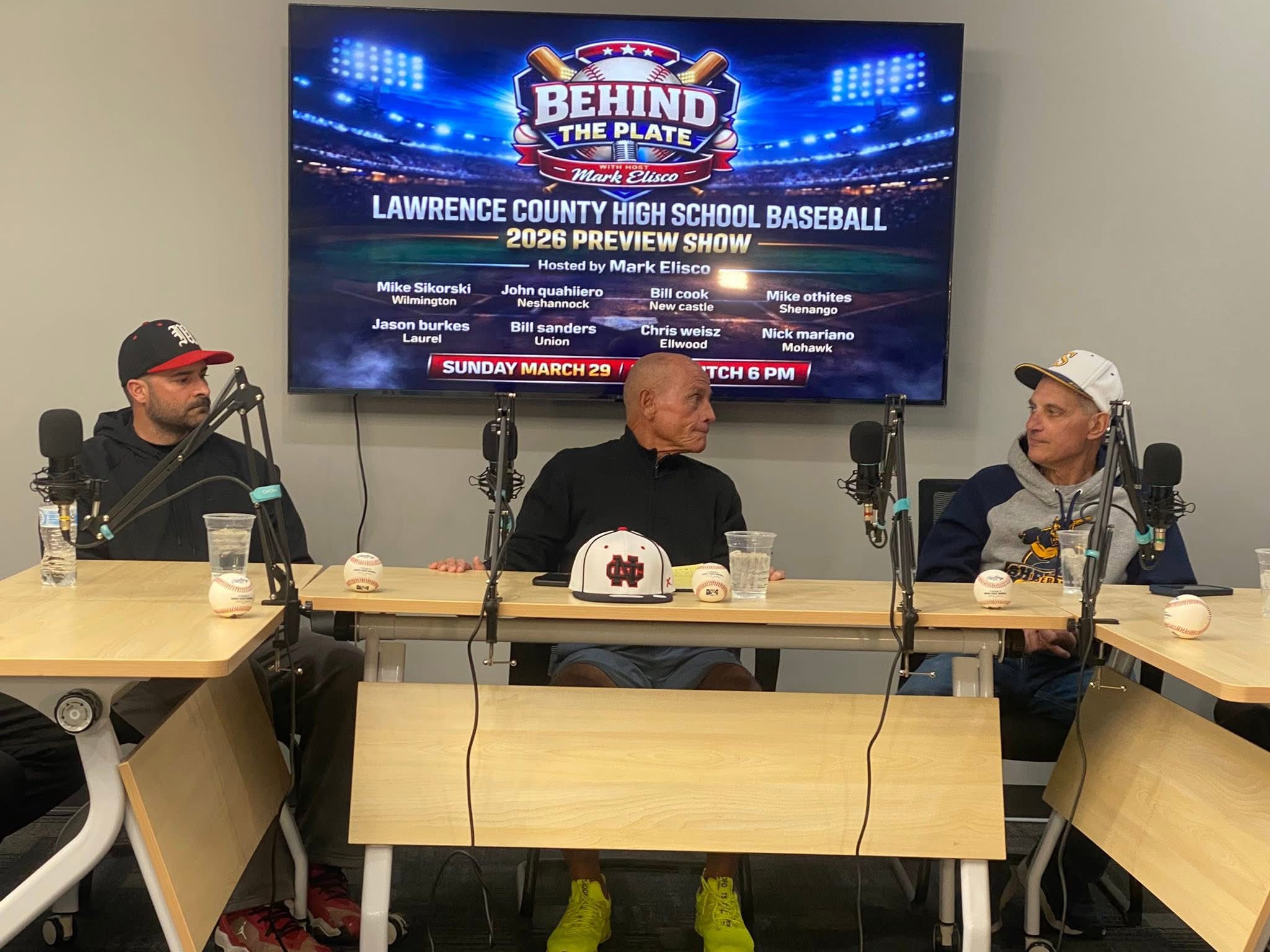 Three people sit at a table with microphones, in front of a large screen displaying a baseball event poster, in a room with beige walls.