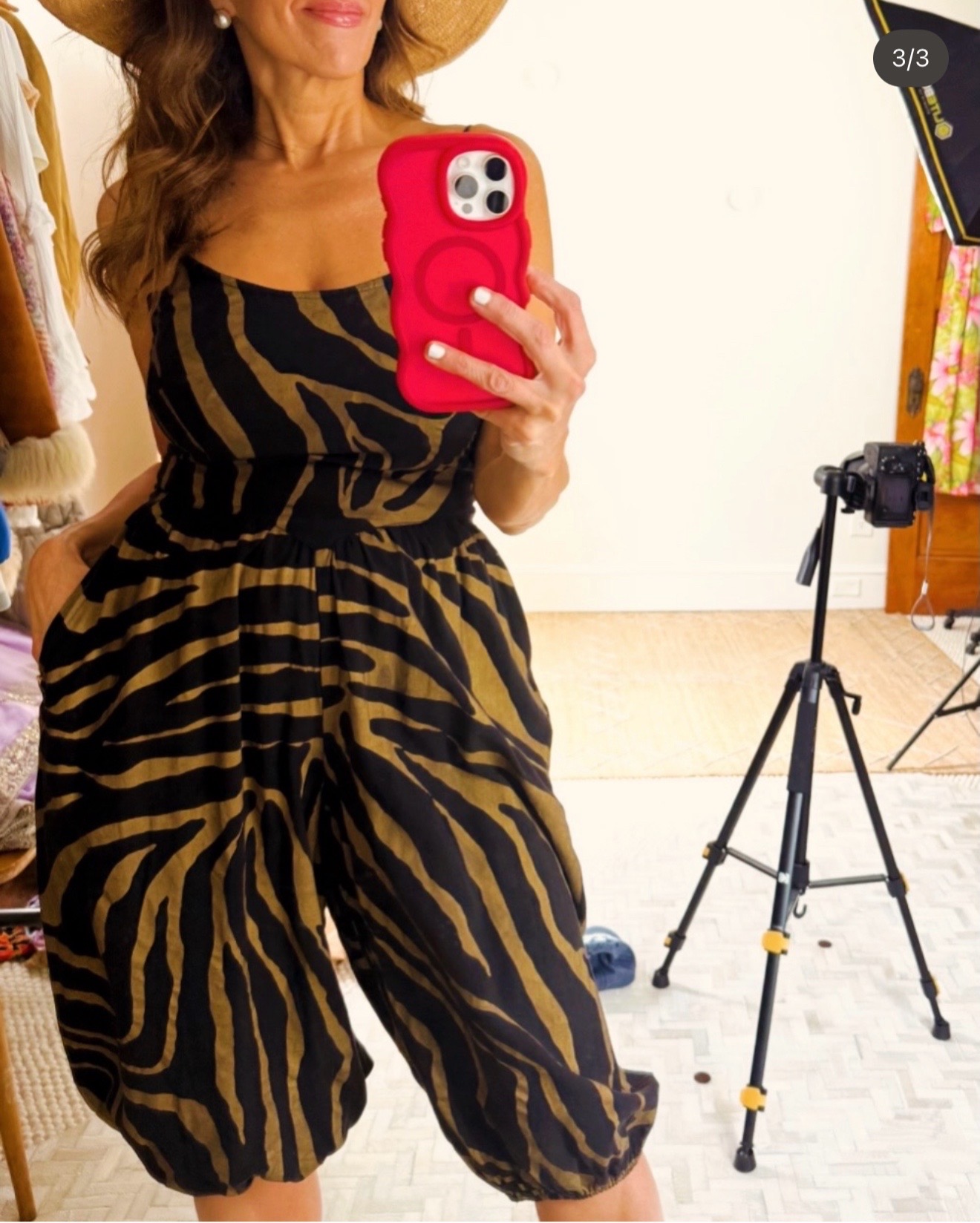 Woman taking a mirror selfie with a red phone, wearing a black and yellow animal print dress, standing in a room with a camera on a tripod.