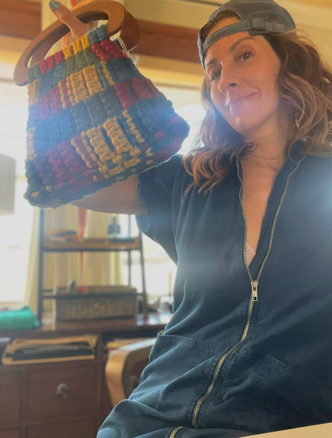 Woman holding colorful knitted teapot, smiling, indoors with shelves and furniture in background.