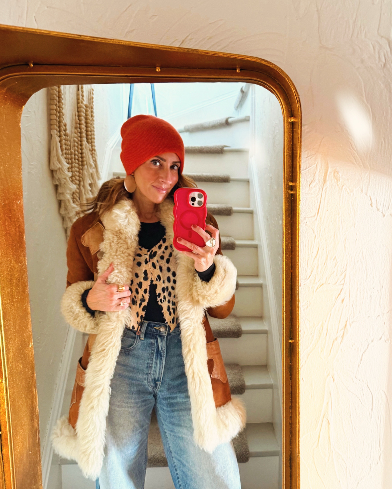 Young woman taking a mirror selfie indoors, wearing a red beanie, leopard print top, and a fur-lined coat.