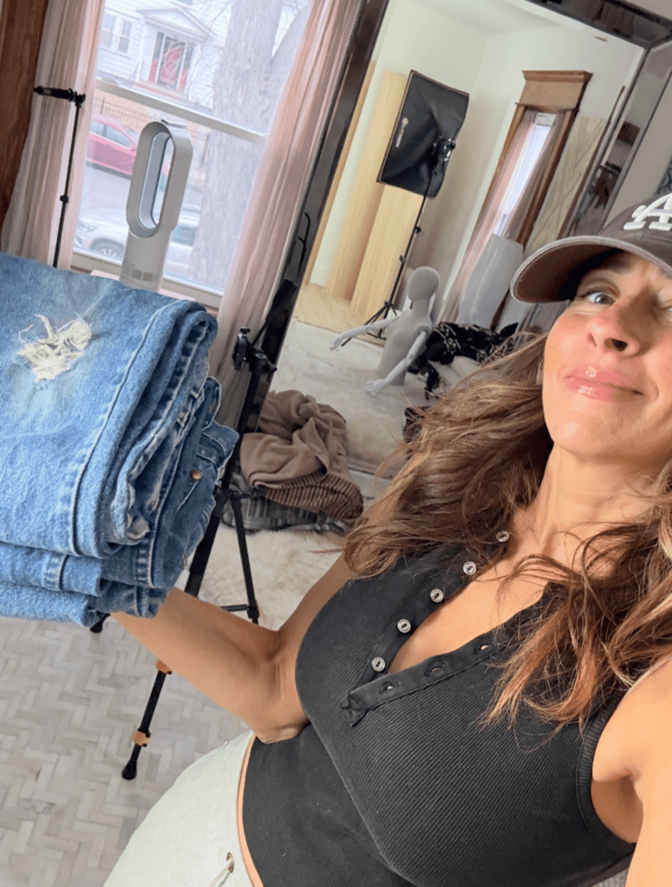 Woman holding a stack of jeans in a room with a dog and a mirror, sunlight coming through window.
