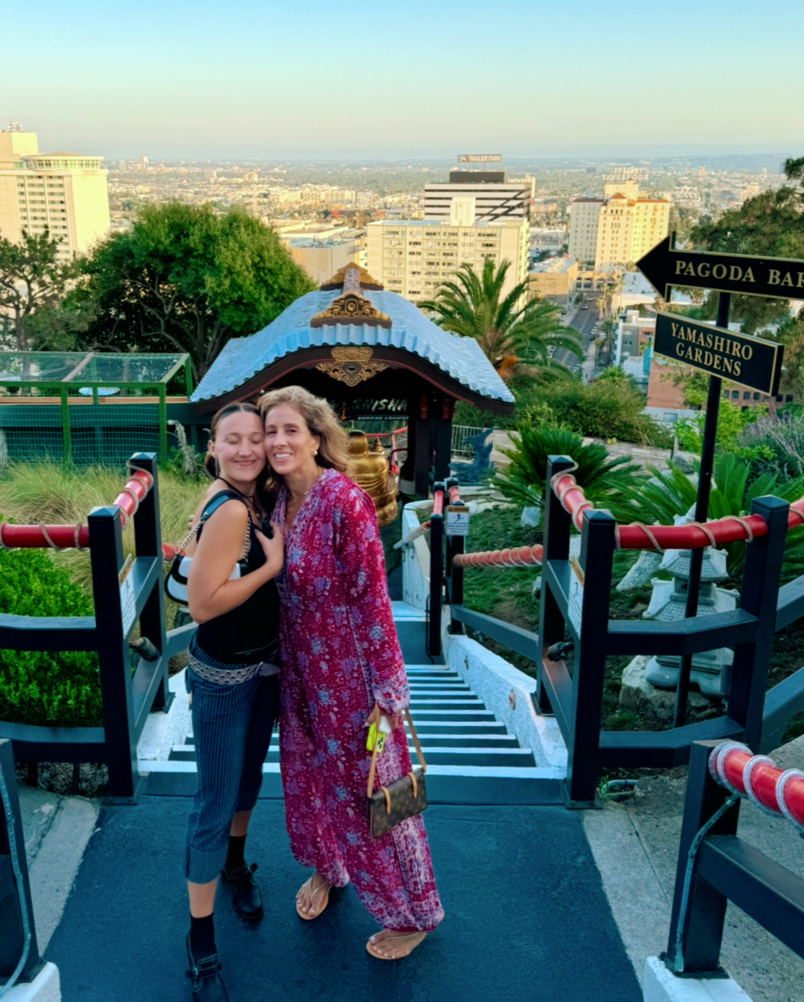 Two women stand on stairs with cityscape background, smiling, one in a black top and jeans, the other in a pink patterned dress.