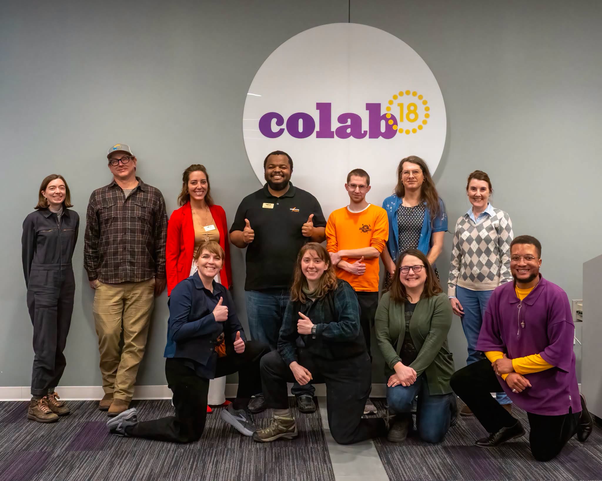 Group of ten people standing and kneeling in front of a wall with a large logo reading 'colab' and '18'.
