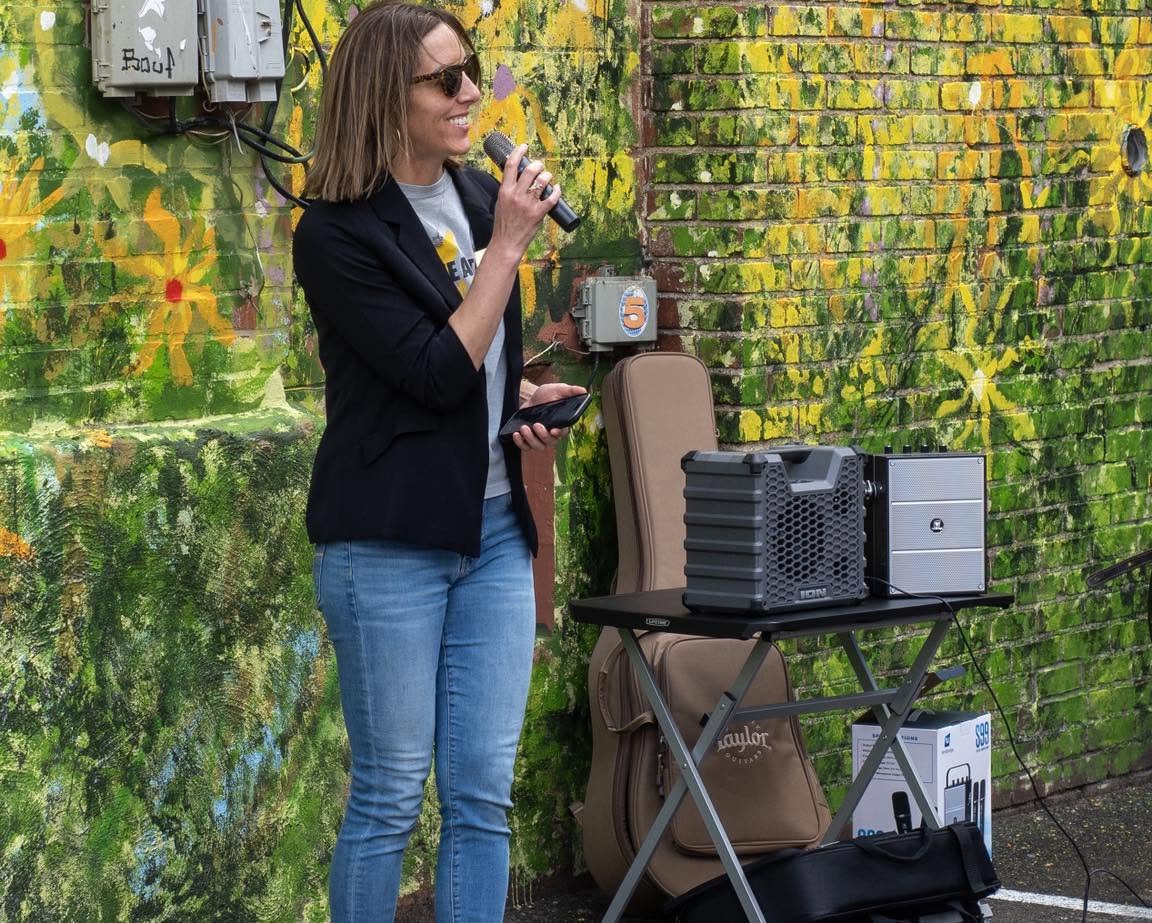 Woman with sunglasses speaking into a microphone outdoors, standing next to a table with audio equipment, against a colorful mural wall.