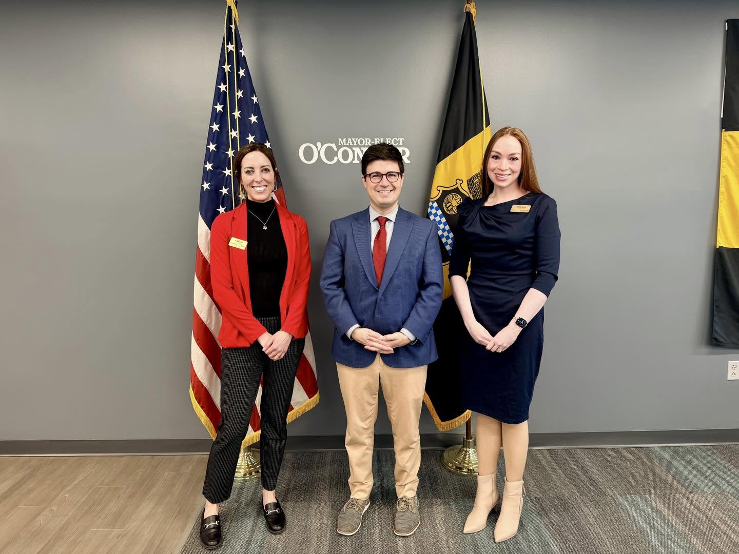 Three people standing in front of American and German flags, smiling, in a professional setting.