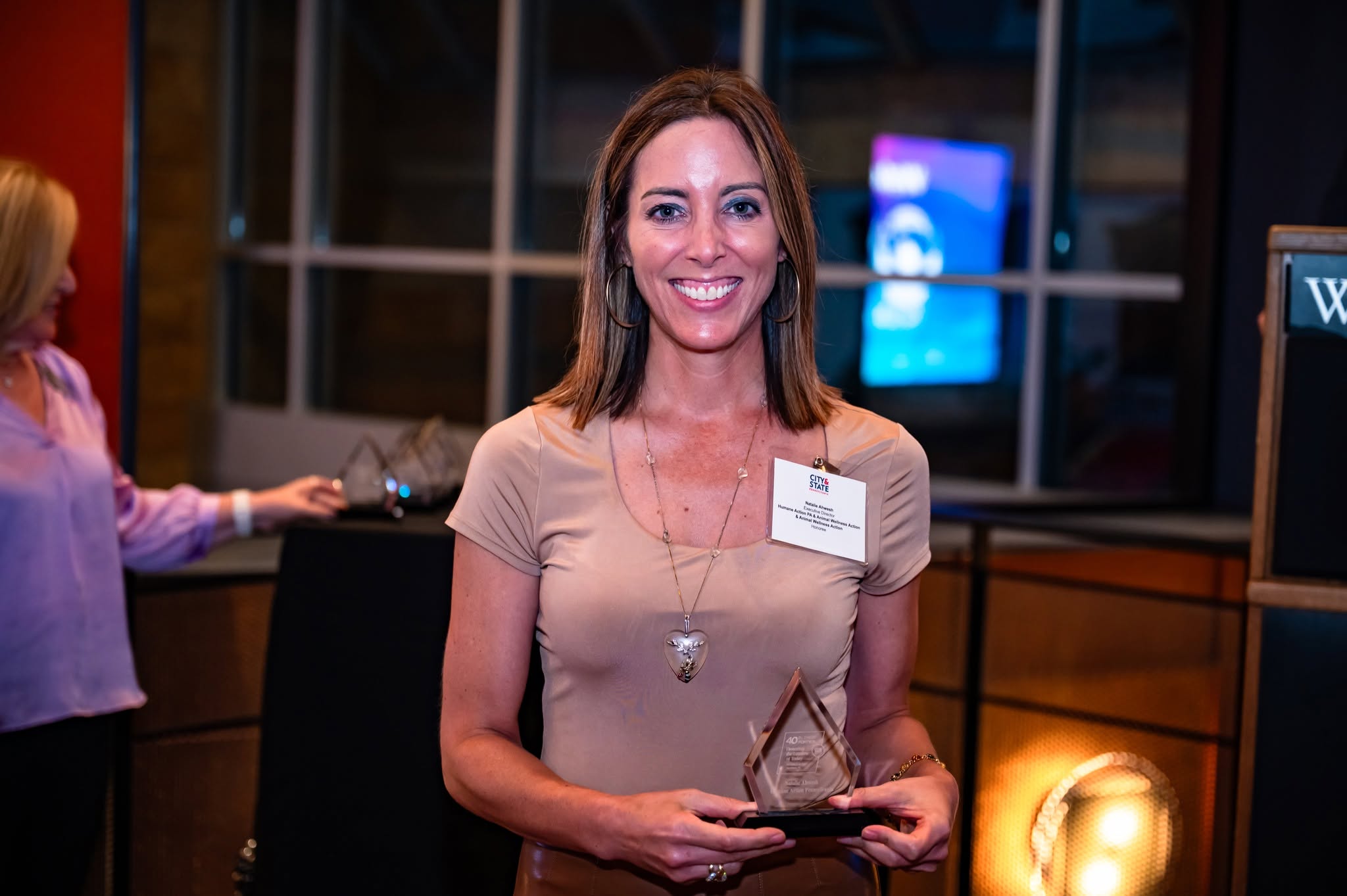 Woman holding an award, smiling, wearing a beige top and necklace, at an indoor event with people and screens in background.