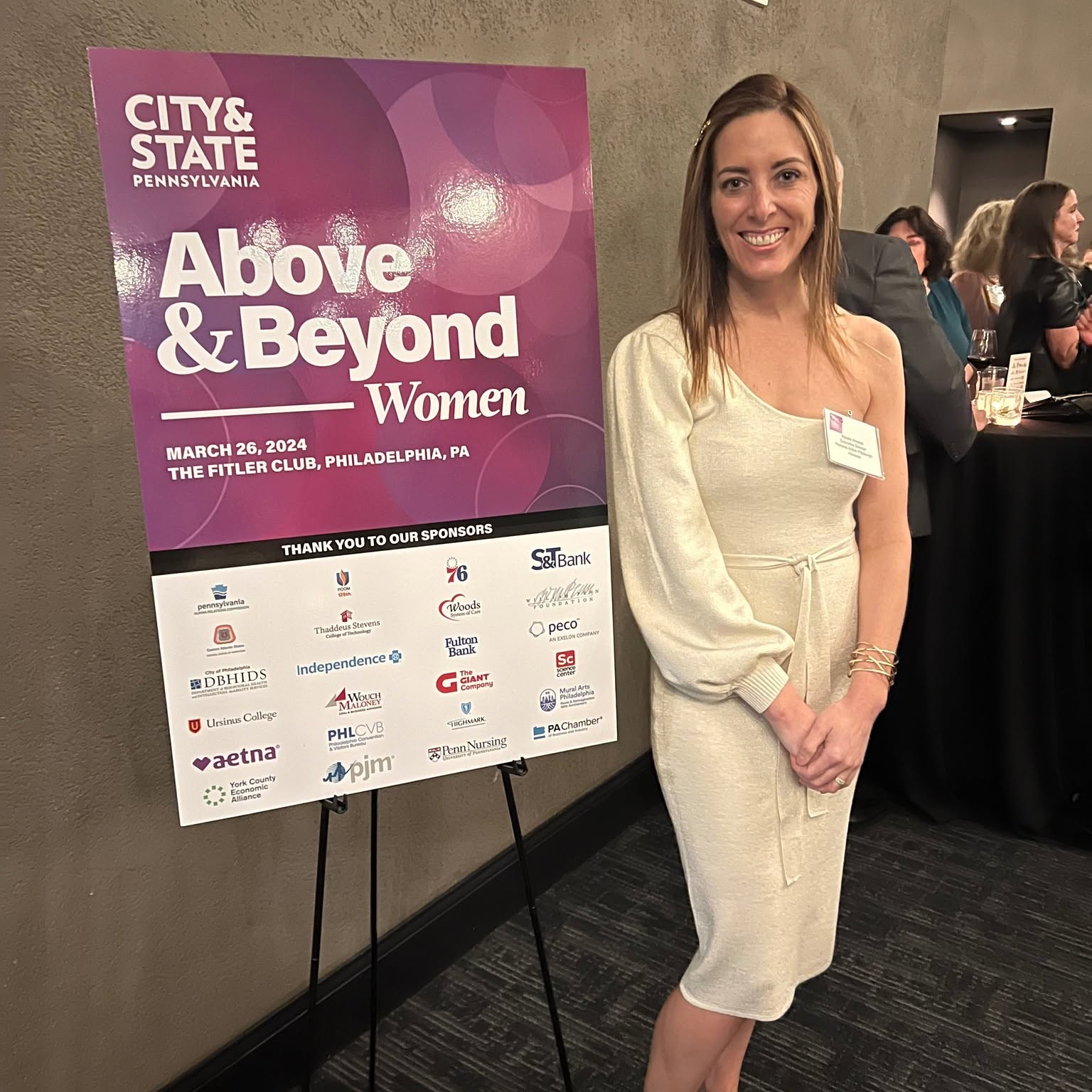 Woman in cream dress standing next to a pink and white event poster at a conference, smiling. Word count: 20.