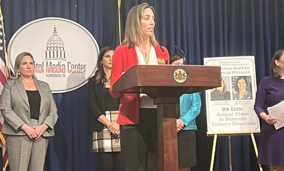Woman in red blazer speaking at a podium during a formal event, with a group of women behind her and a sign on the right.