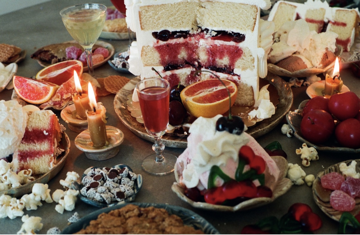 Assorted cakes, desserts, candles, and fruits on a table, with a layered cake and a decorated cupcake visible.
