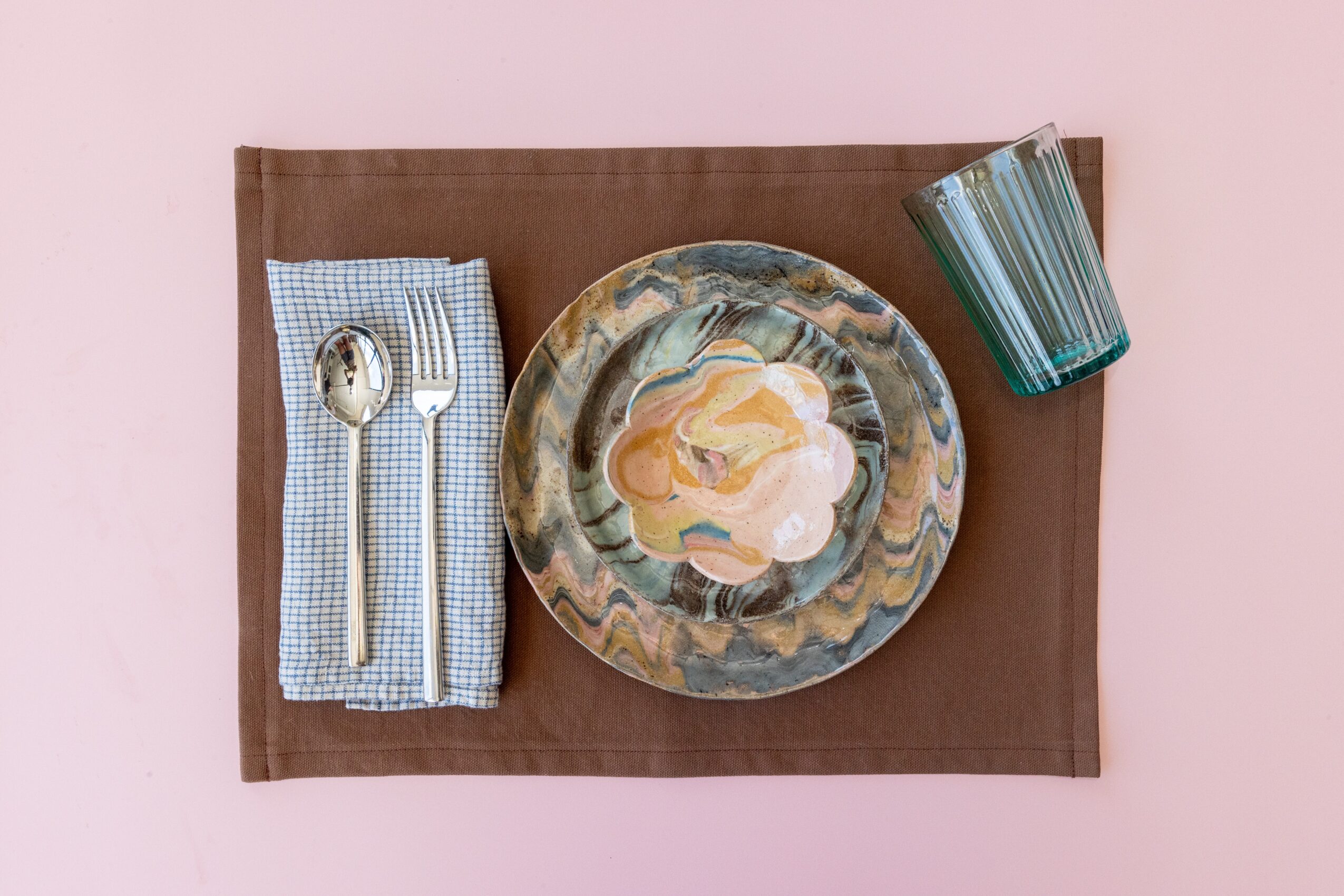 Place setting with a plate, spoon, fork, glass, and napkin on a pink background.