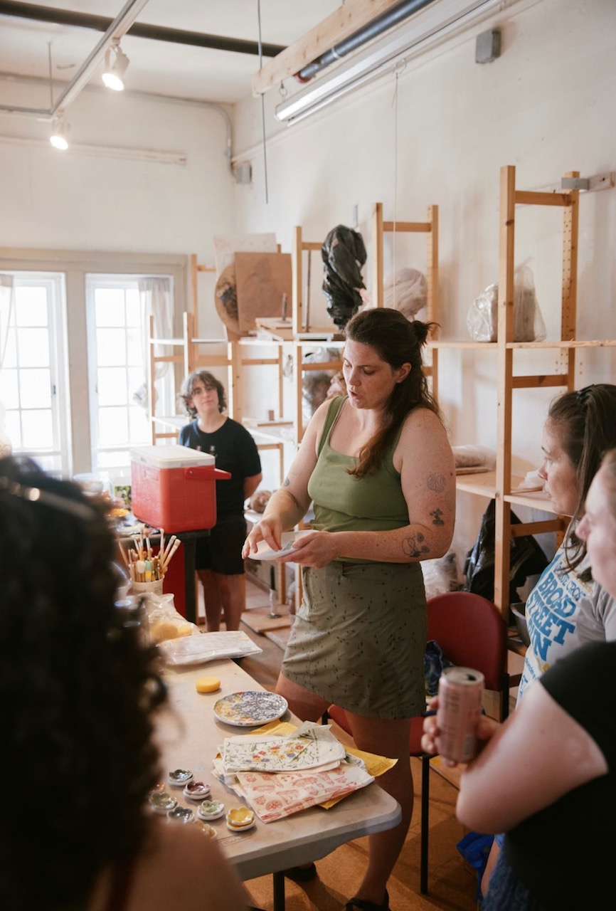 Woman standing at table with food, surrounded by people in a bright room with shelves and windows.