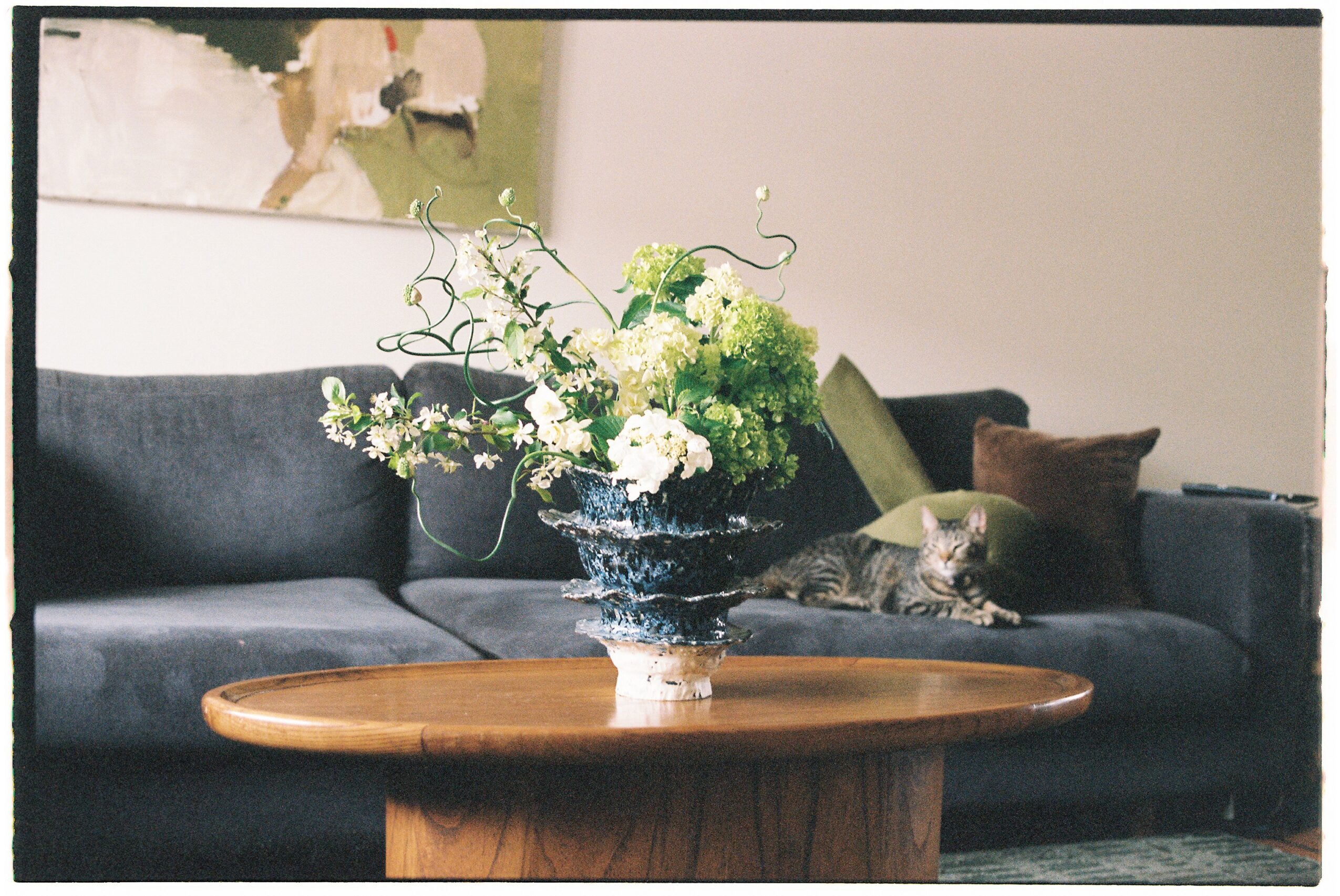 Flower arrangement on a wooden table in front of a gray sofa with pillows and a cat resting on it.