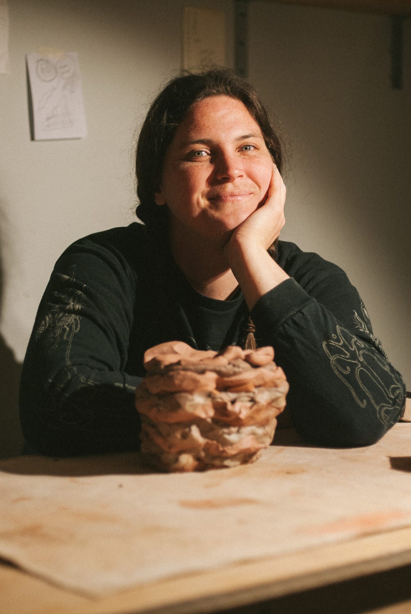 Person with short dark hair smiling, resting face on hand, sitting at table with a stack of cookies, indoor setting.