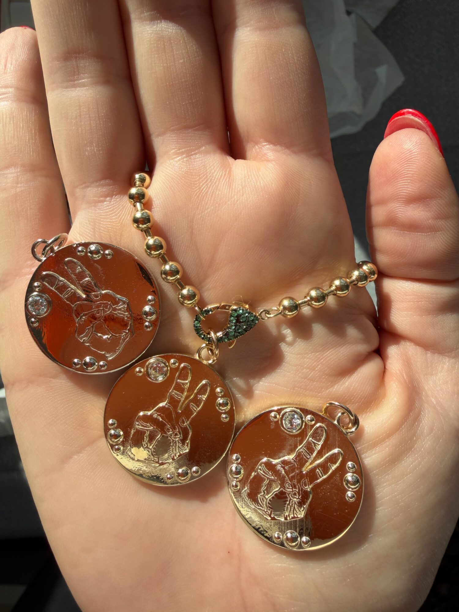 Hand holding a necklace with three round, gold-colored pendants featuring a raised figure and small decorative elements.