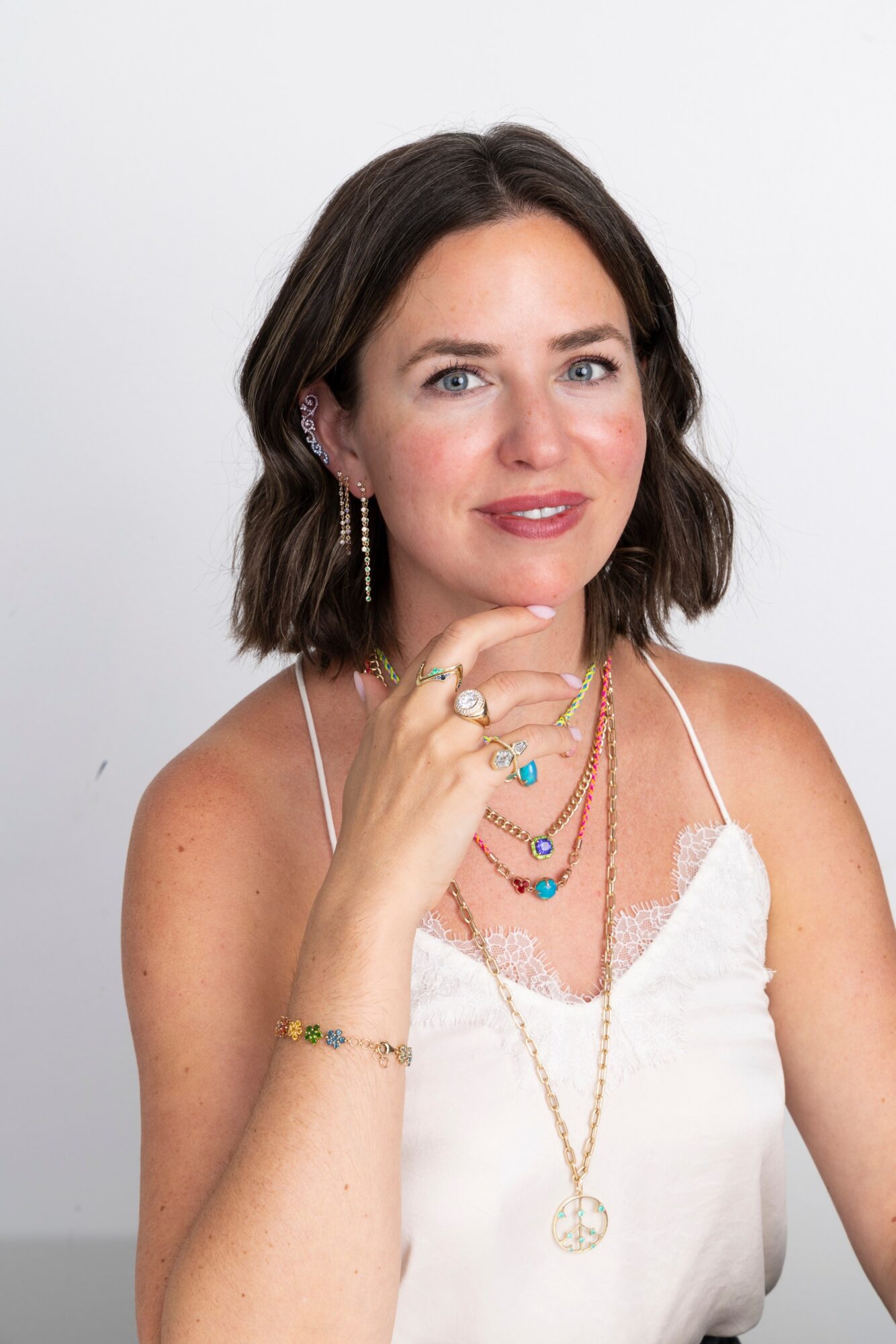 Woman with shoulder-length dark hair, wearing a white top and layered jewelry, smiling and touching her chin.