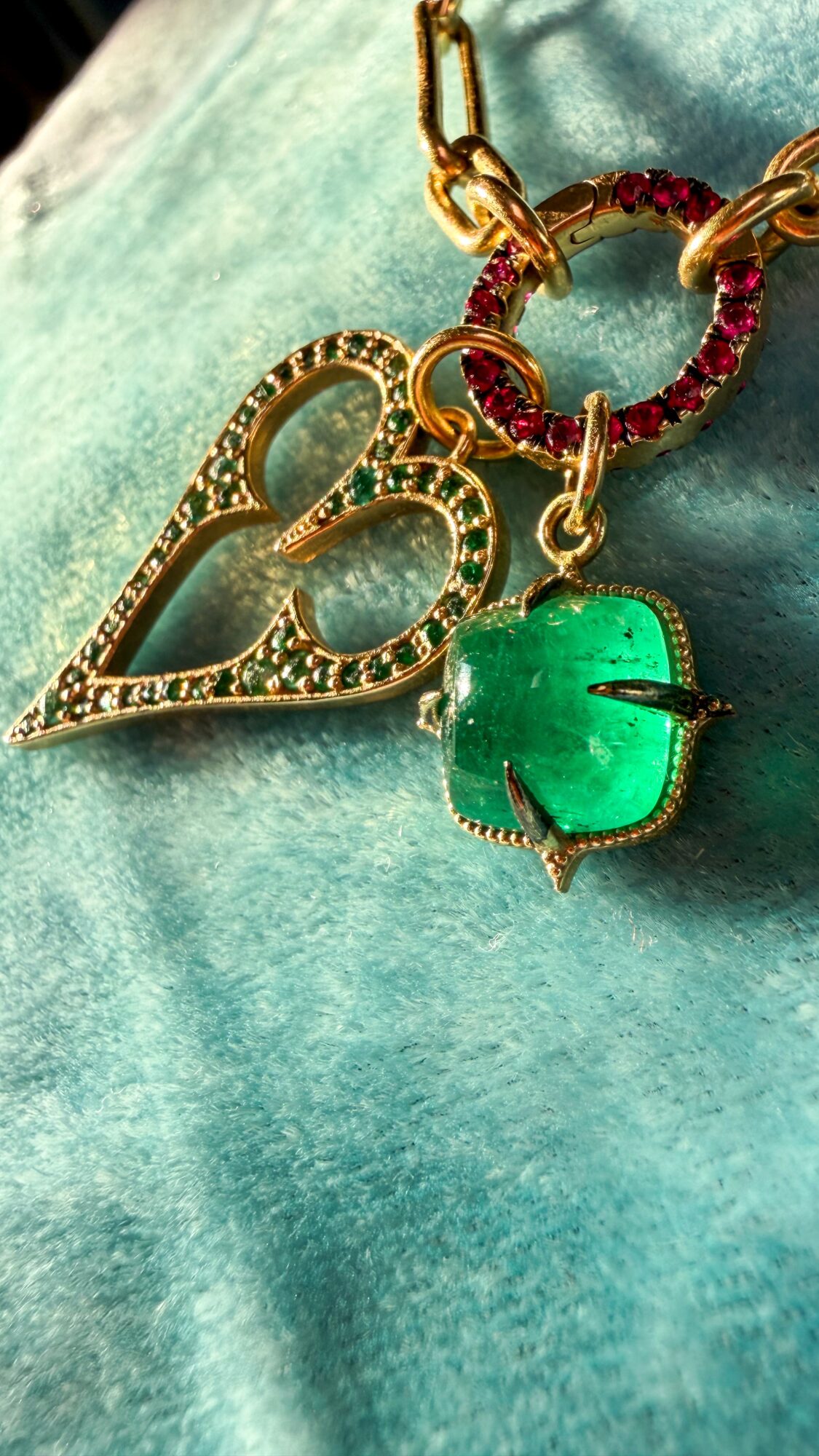 Close-up of a gold heart-shaped pendant with rhinestones, a green gemstone, and a red beaded ring on a teal fabric background.