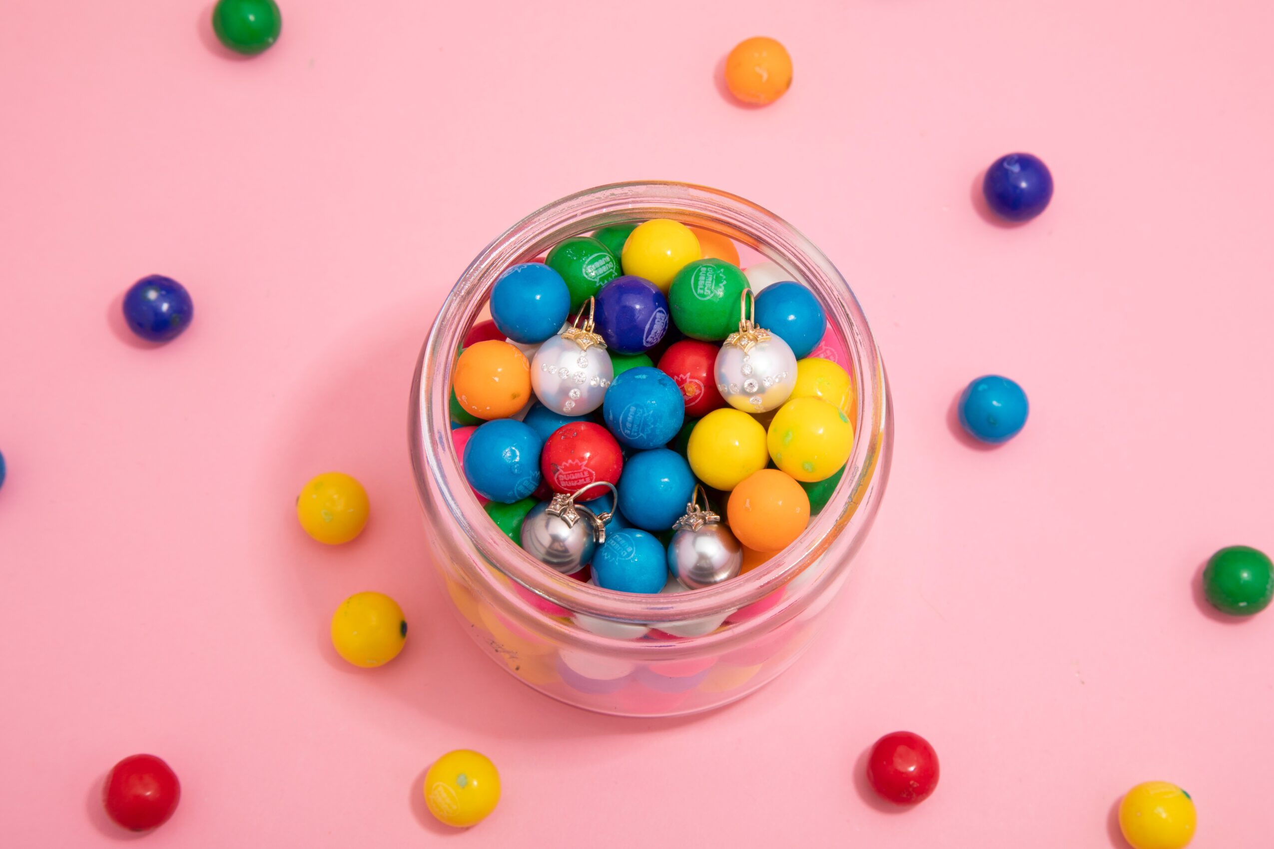 Colorful gumballs in a glass jar on a pink background with scattered gumballs around it.