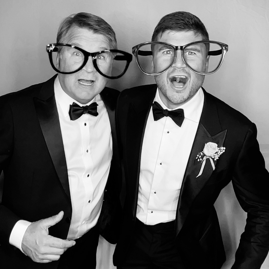 Two men in tuxedos wearing large glasses, posing playfully, one with a bow tie and boutonniere, black and white photo.
