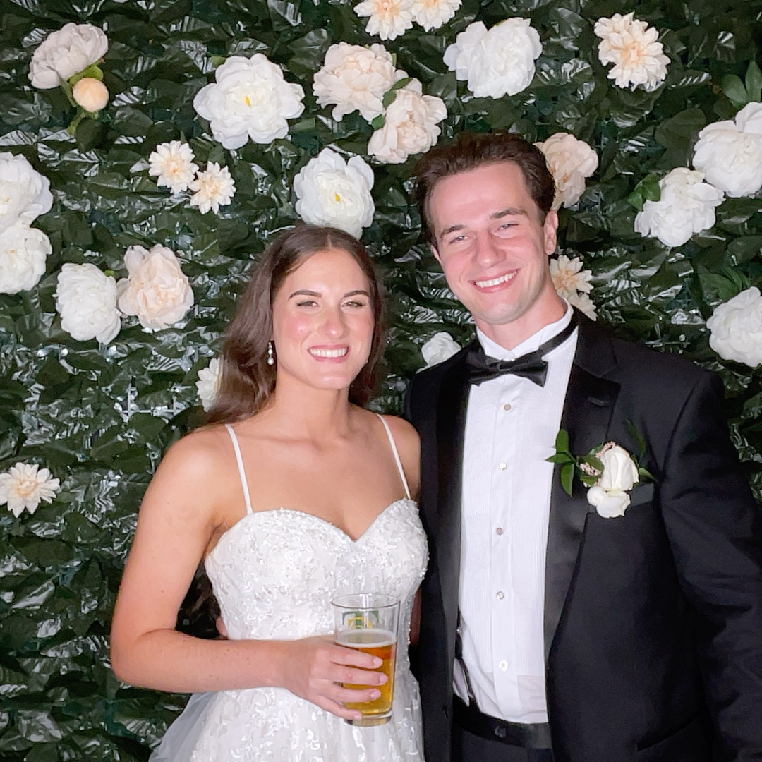 A smiling couple in formal attire standing in front of a floral backdrop, the woman holding a drink.