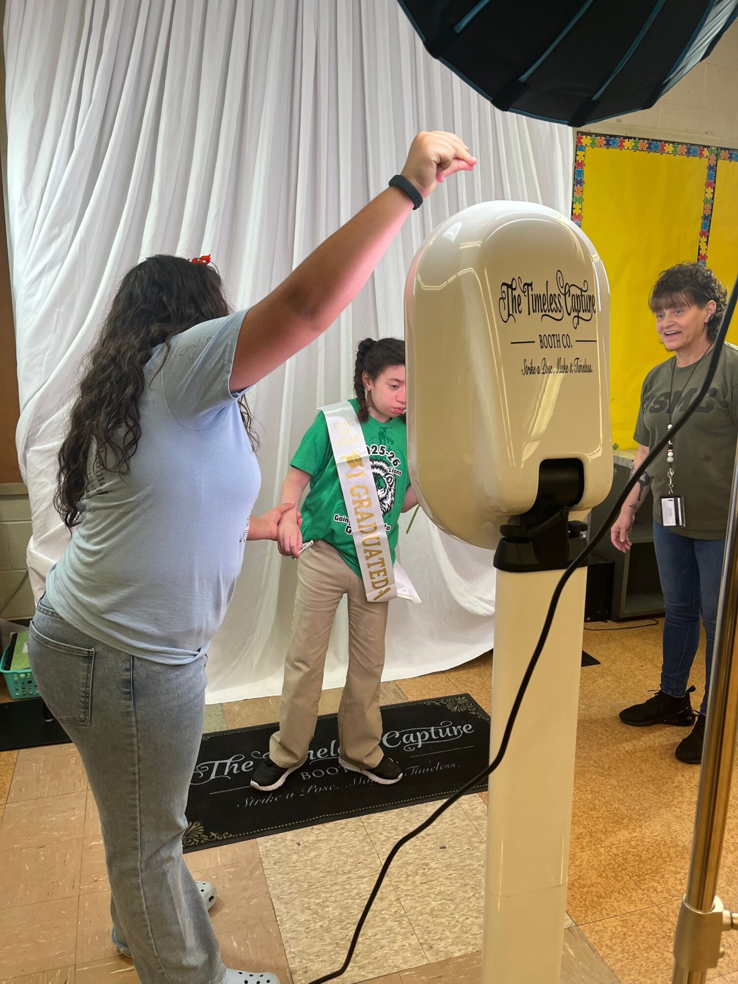 Person with long dark hair speaking into a microphone, standing near a large white object, with a person in a green jacket in the background.