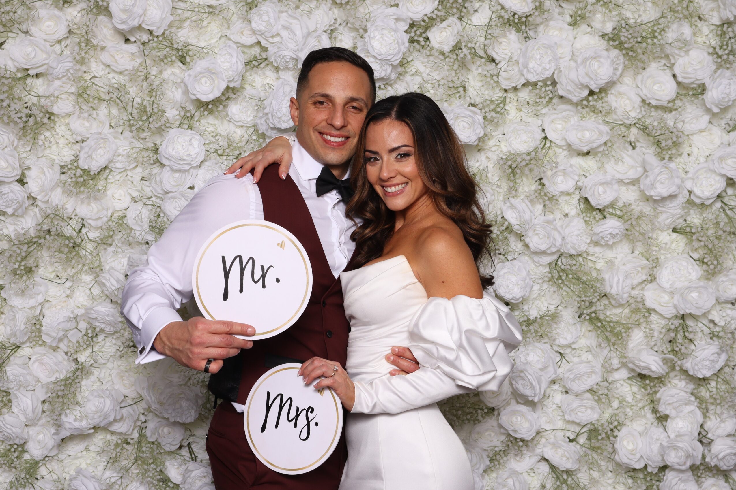 A smiling couple dressed in formal attire holding signs that say 'Mr.' and 'Mrs.' in front of a floral backdrop.