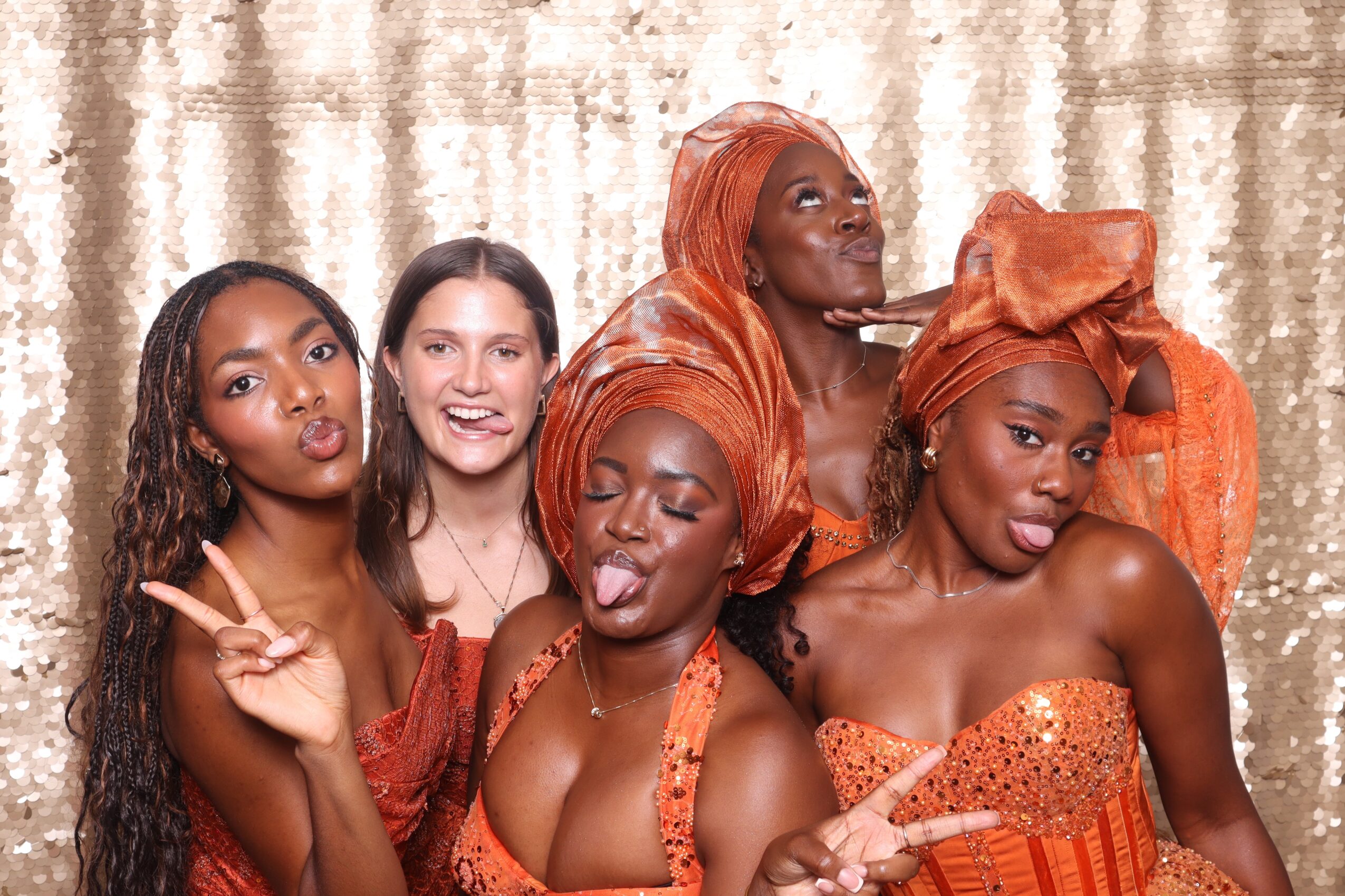 Five women posing together, some making peace signs and sticking out tongues, in front of a gold curtain backdrop.