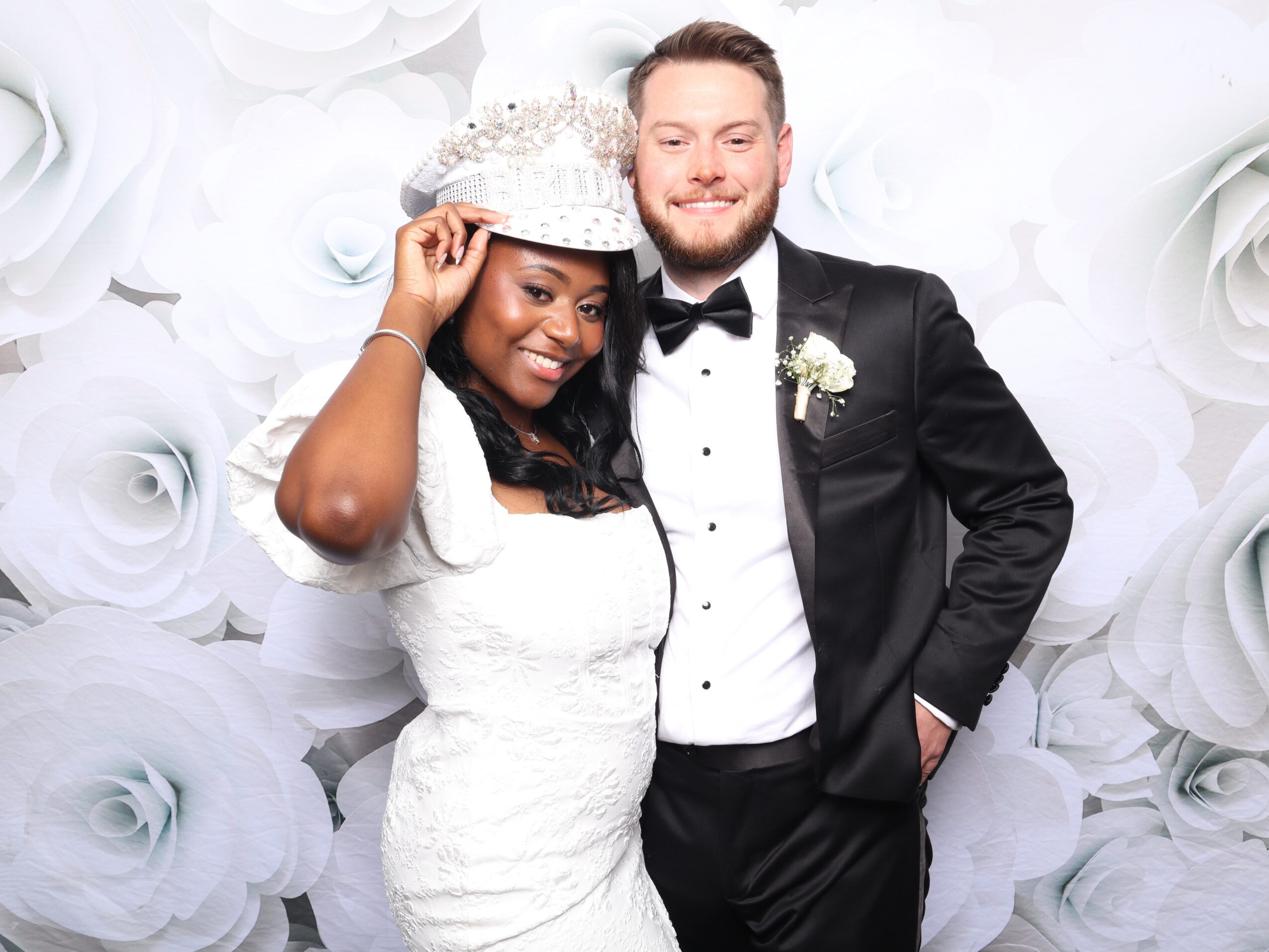 A bride and groom pose together, the bride wearing a white dress and crown, the groom in a tuxedo, smiling.