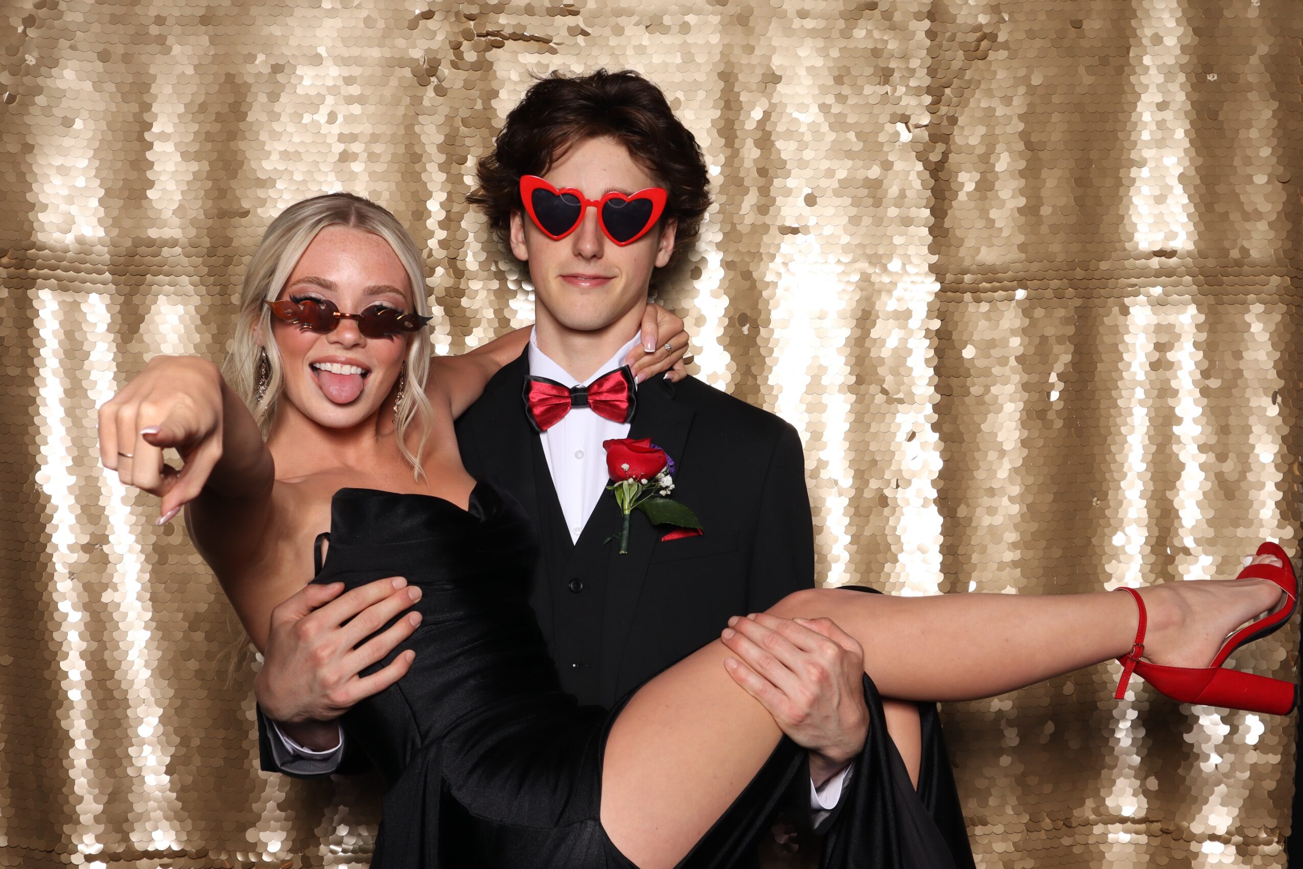 Two people dressed in formal attire, one holding the other horizontally, with a gold sequin backdrop.