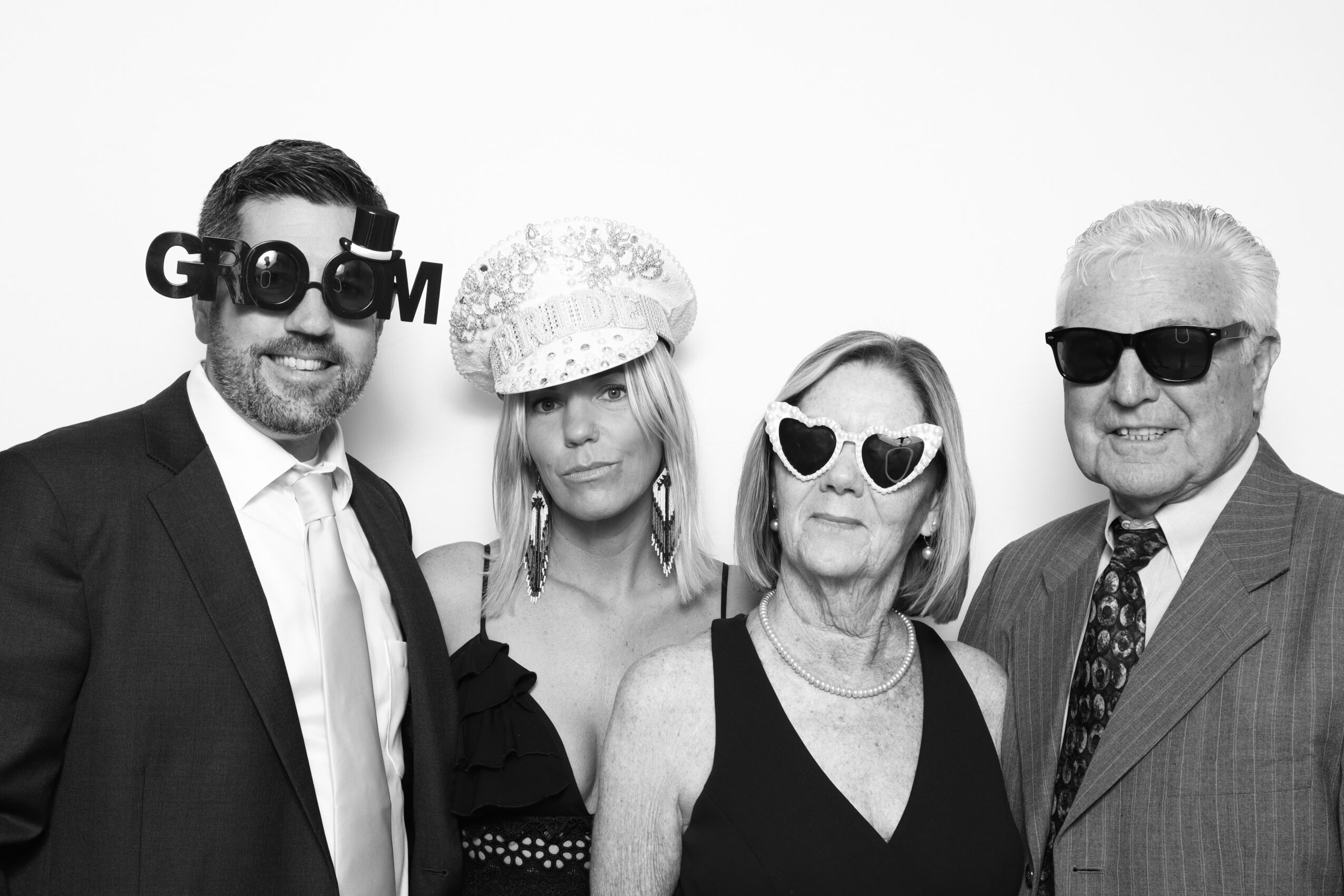 Four people in formal attire wearing sunglasses, standing together against a plain background.