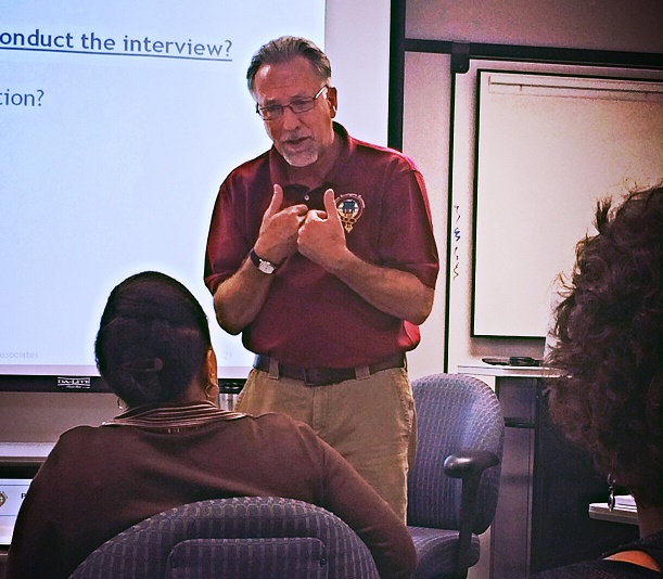 Man in red shirt speaking to a group in a classroom, with a presentation slide behind him.