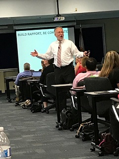 Man in white shirt and tie standing in front of classroom, gesturing with arms open, students seated at desks, presentation screen behind.