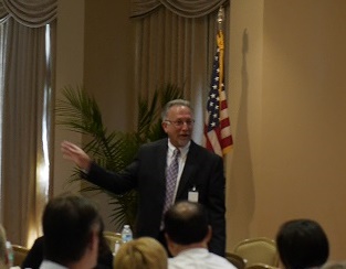 Man in suit speaking at a conference with an American flag and plants in the background.