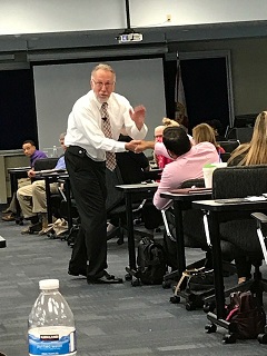 Man standing and speaking to seated group in classroom, with a projector screen behind him.