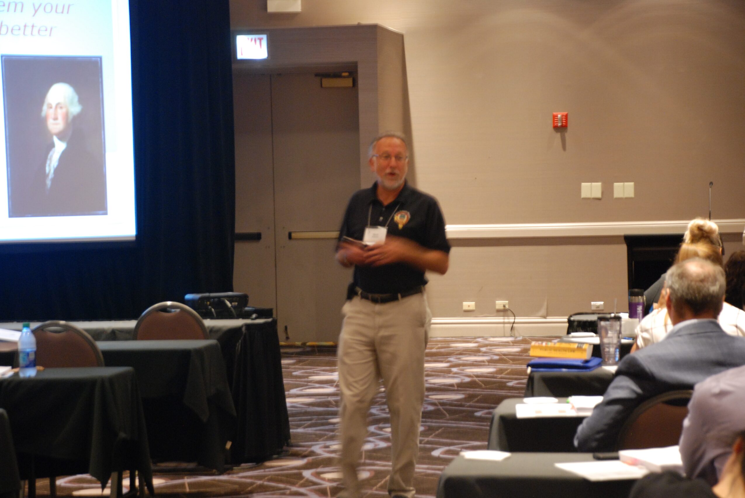 Man standing and speaking in a conference room with seated audience, presentation screen on the left, and tables with materials.