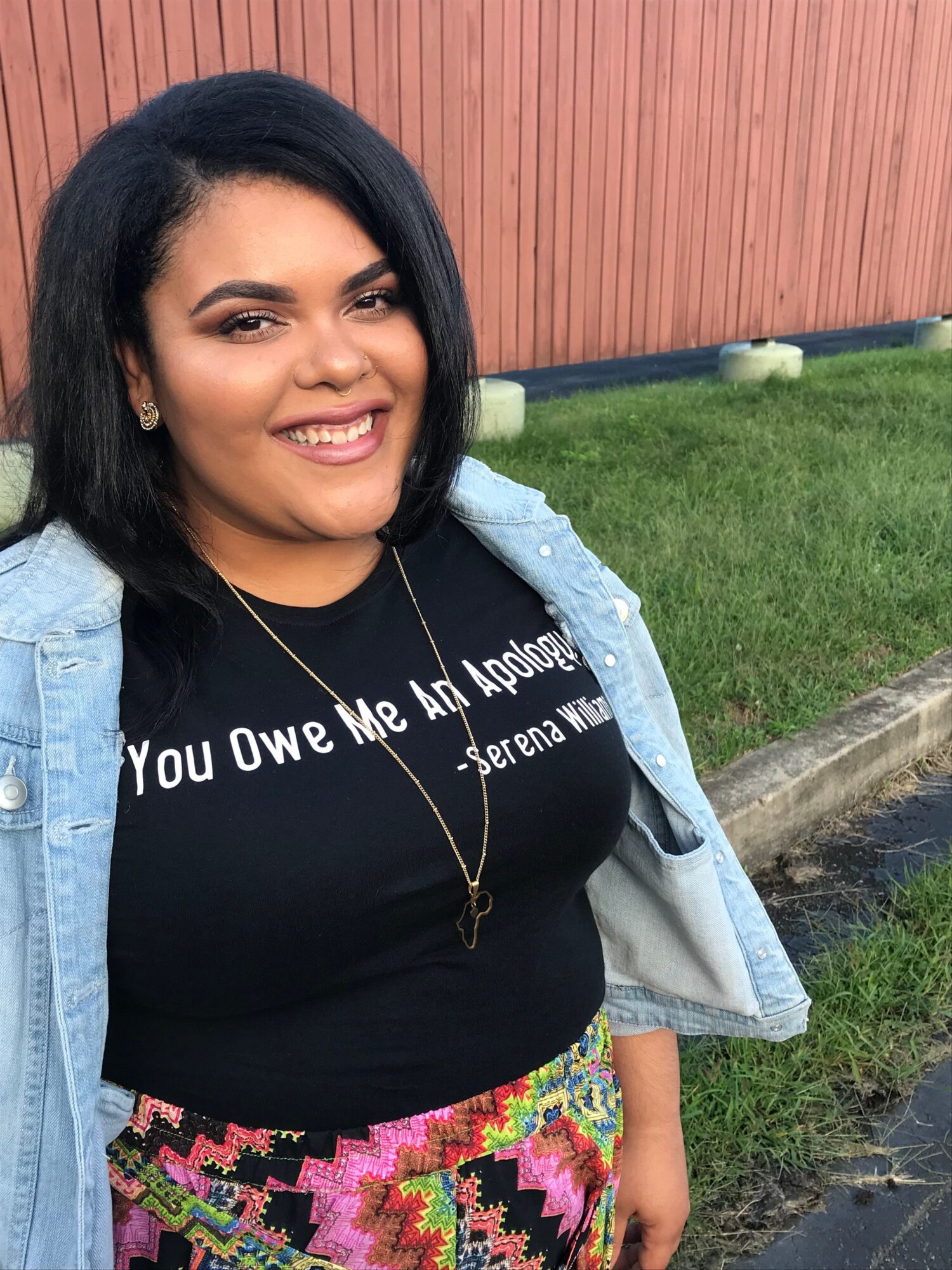 Smiling woman with black hair, wearing a black shirt with white text, a light blue jacket, and colorful patterned pants, outdoors.