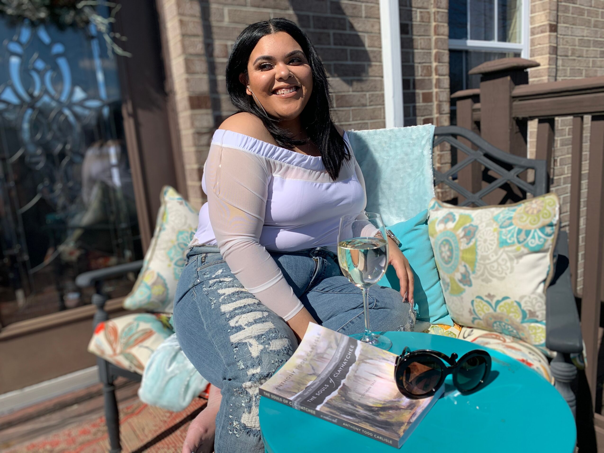 Smiling woman sitting outdoors on a bench with pillows, holding a glass of wine, with a book and sunglasses nearby.