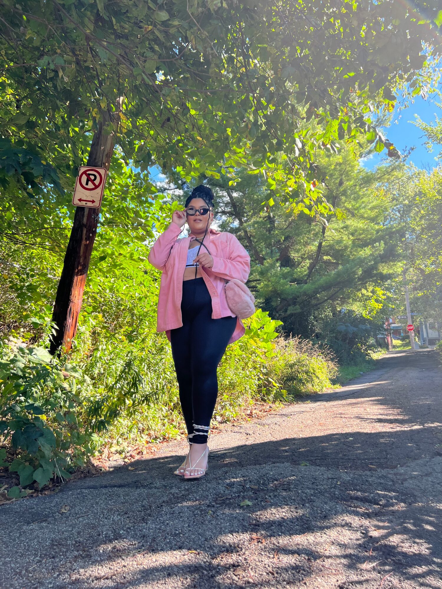 Woman standing on a dirt path surrounded by trees and greenery, wearing sunglasses, pink jacket, and dark pants.