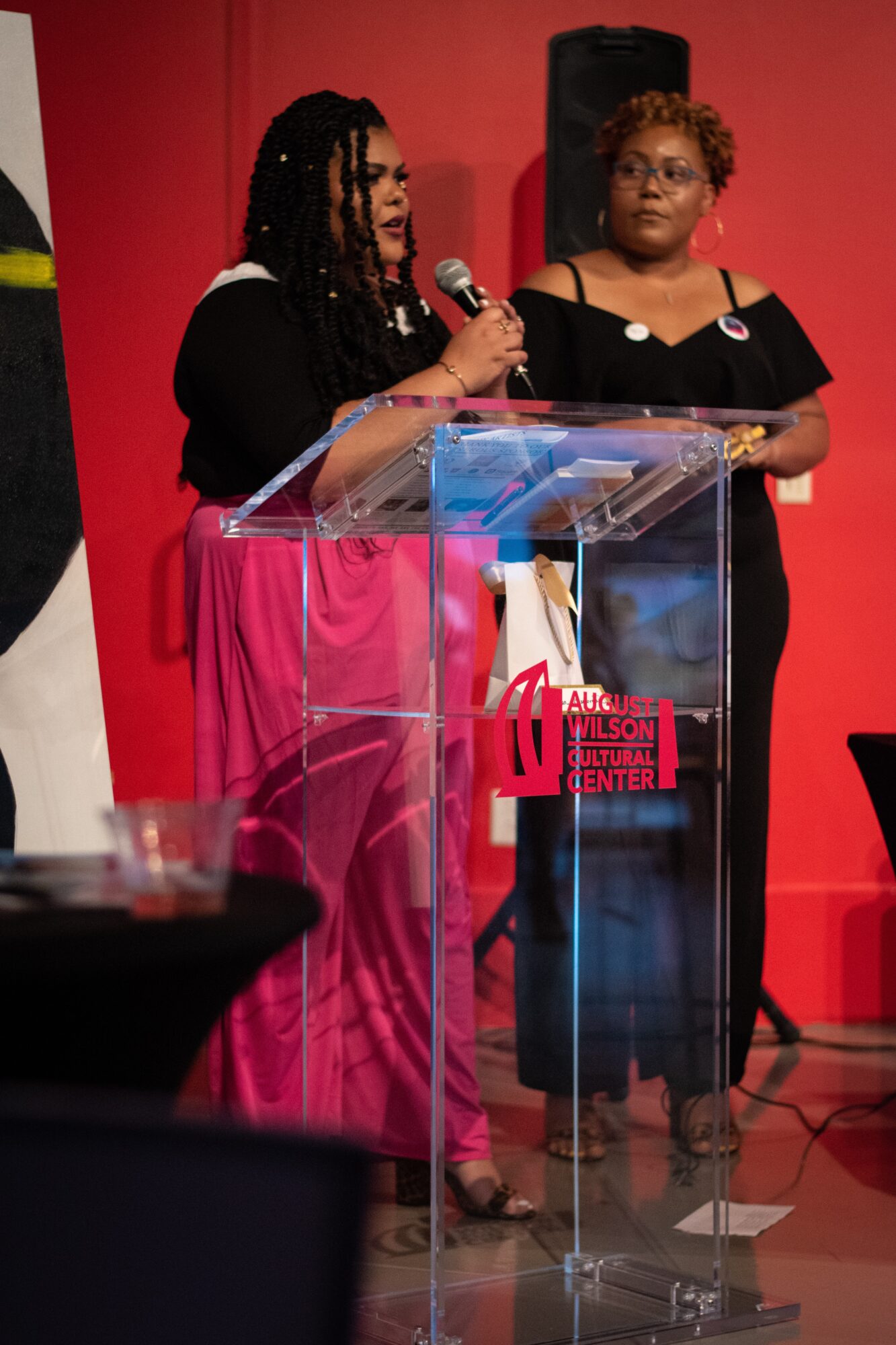 Two women stand at a clear podium with a microphone, speaking at an event with a red background.