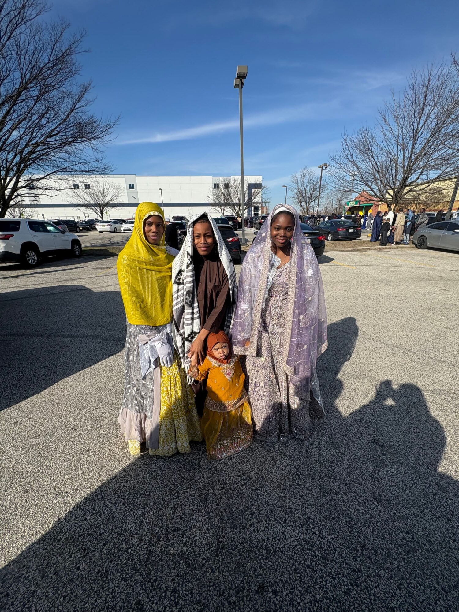 Three women standing outdoors in a parking lot, with trees and a building in the background, under a partly cloudy sky.